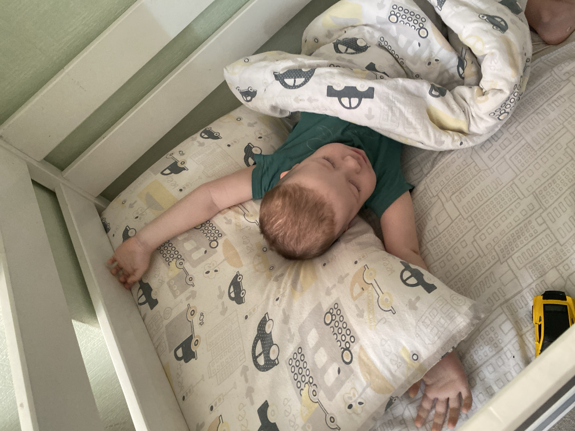 A child is sleeping in a crib. The child is lying on a pillow and under a blanket with car designs. The crib sheets also have a car design. The child is wearing a green shirt. A yellow toy car is visible near the edge of the crib. The image does not provide sufficient information to identify a location in Sochi, Russia.