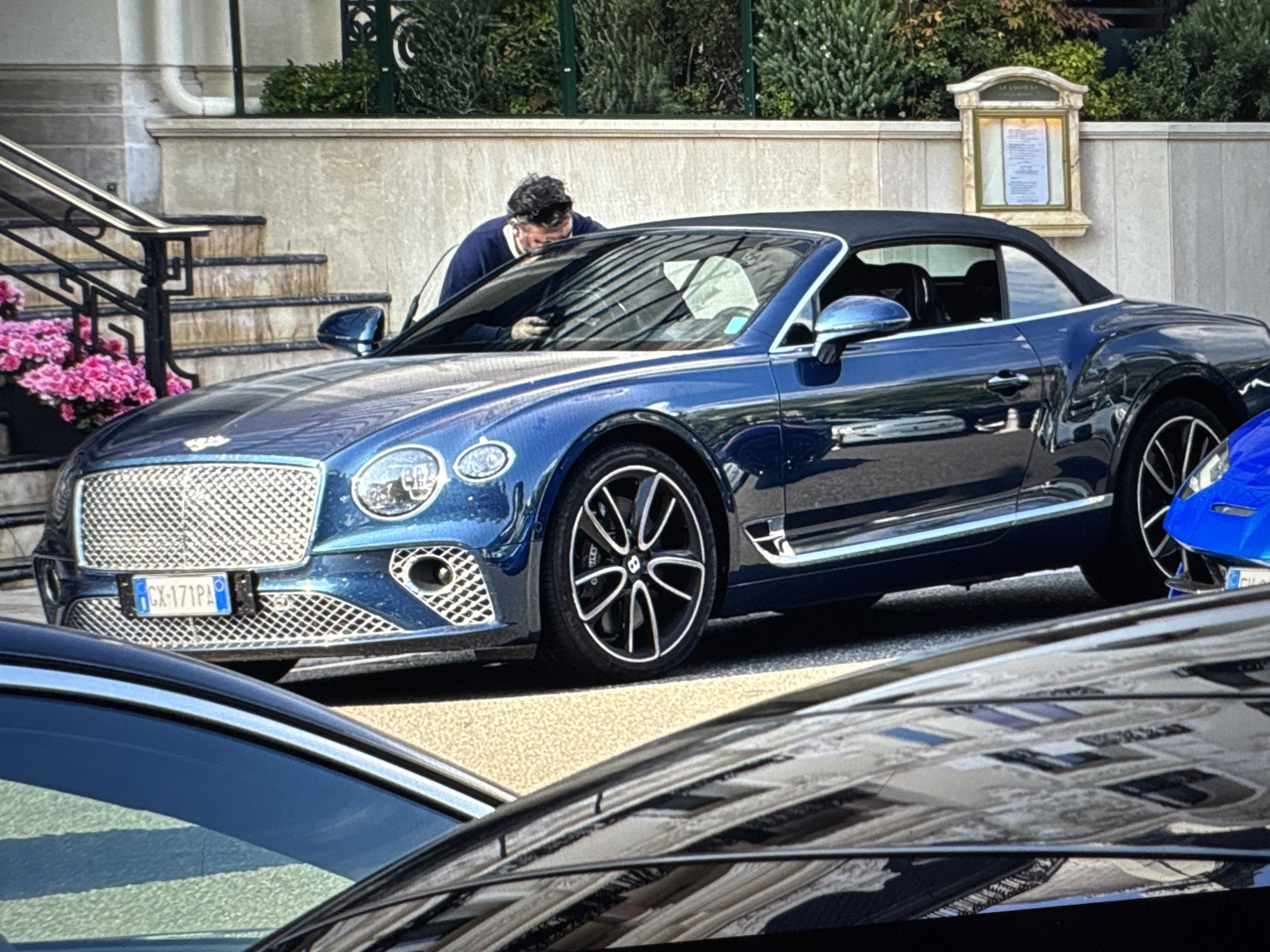 A man, wearing a dark blue sweater and light-colored pants, is bent over the windshield of a dark blue convertible Bentley, looking at his phone. The car is parked on a street, with a building facade and lush green foliage in the background. In front of the building, there are stairs with pink flowers in a planter to the left. The license plate of the Bentley reads "CX-171PA". The scene appears to be in an urban setting.