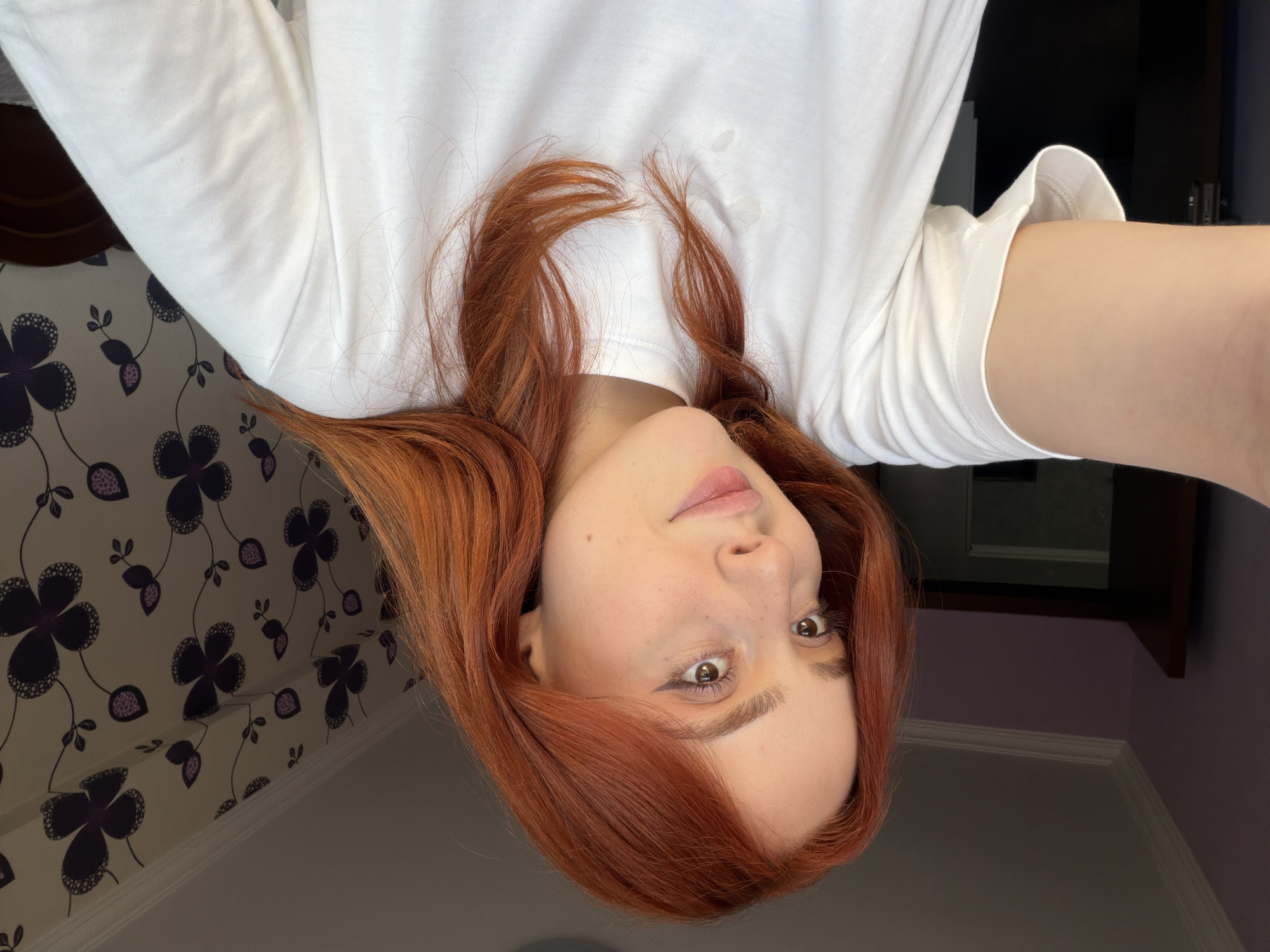 A young woman with red hair is taking a selfie. She is wearing a white long-sleeved shirt. The background includes a wall with a floral pattern (dark purple flowers on a light background) and a dark-colored door. The location is not discernible from the image. The image's orientation is rotated.