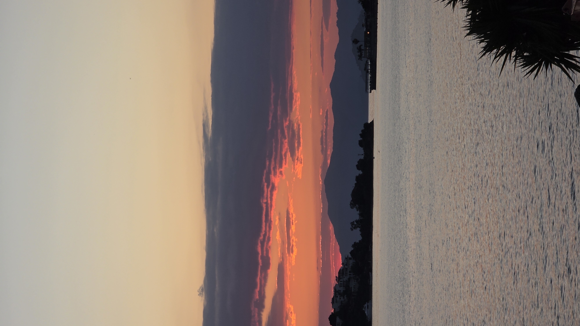 The image depicts a sunset over the water, possibly in Fethiye, Turkey, given the mountain silhouettes in the background. The sky transitions from light yellow near the top of the image to a vivid orange and red closer to the horizon. Dark, layered clouds are visible. The water surface reflects the colors of the sky. A silhouette of trees is visible on the right and on the horizon, along with a structure that may be a dock or pier.