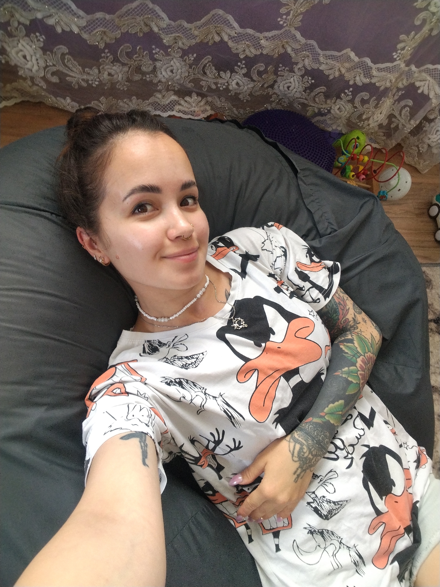 A young woman with her hair in a bun and a nose ring lies on a dark grey beanbag chair. She is wearing a white t-shirt with an all-over print of Daffy Duck characters. She has extensive tattoos on her arms, including one that covers her left arm from the shoulder down. She is also wearing a beaded necklace. In the background, there is a sheer, lace curtain with a floral pattern, and several children's toys. The woman is looking at the camera and smiling.