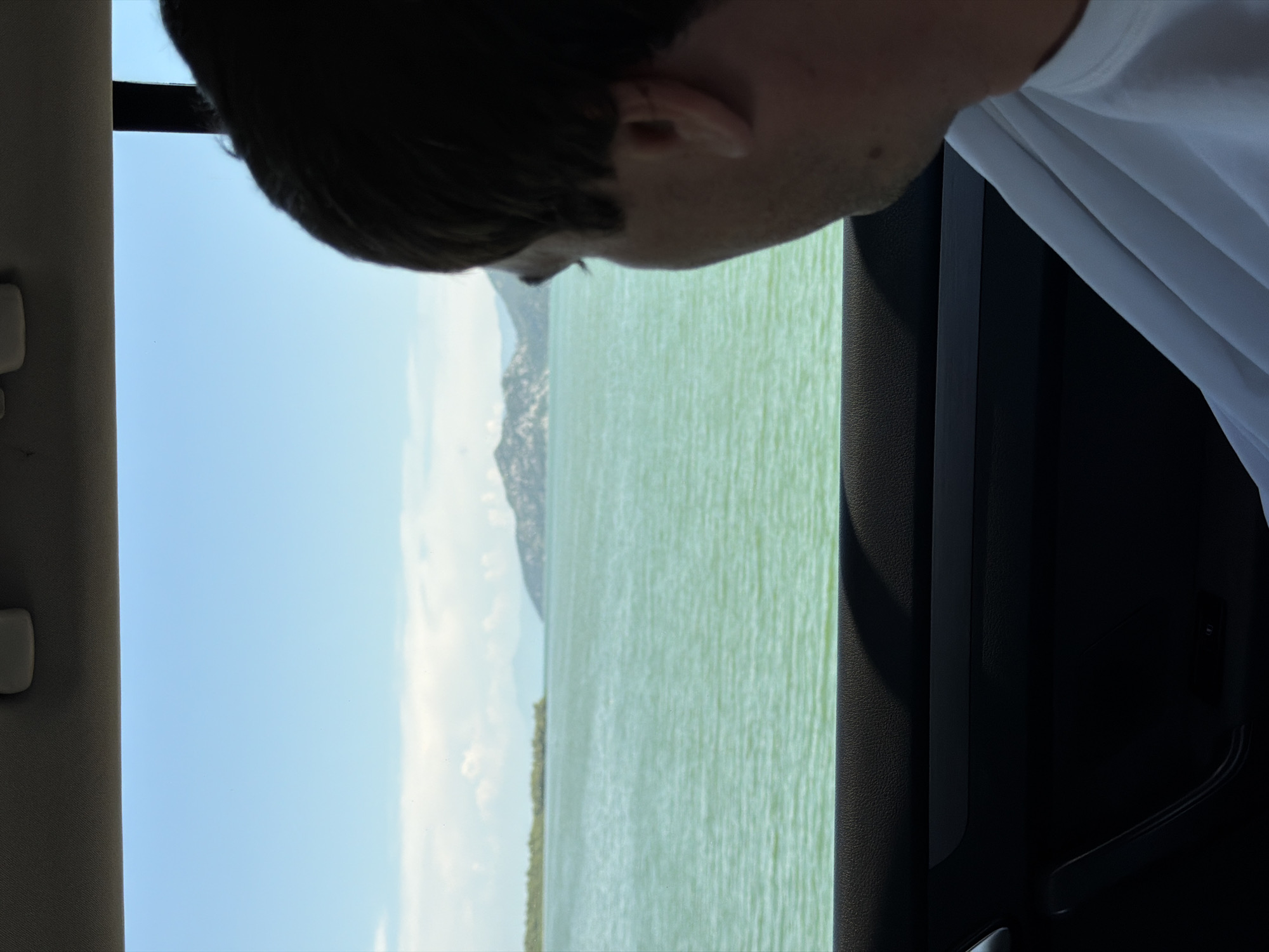 The image, taken from inside a vehicle, captures a view of a wide body of water with distant hills and a bright blue sky. The water is a vibrant green, with small ripples suggesting a gentle breeze. A person's head and shoulder, partially obscured by the frame, are visible in the foreground, looking out the window. The interior of the car is visible at the bottom, with dark-colored upholstery and a door handle. The scene appears to be located in Kruševica, Montenegro, given the characteristic landscape of the region.