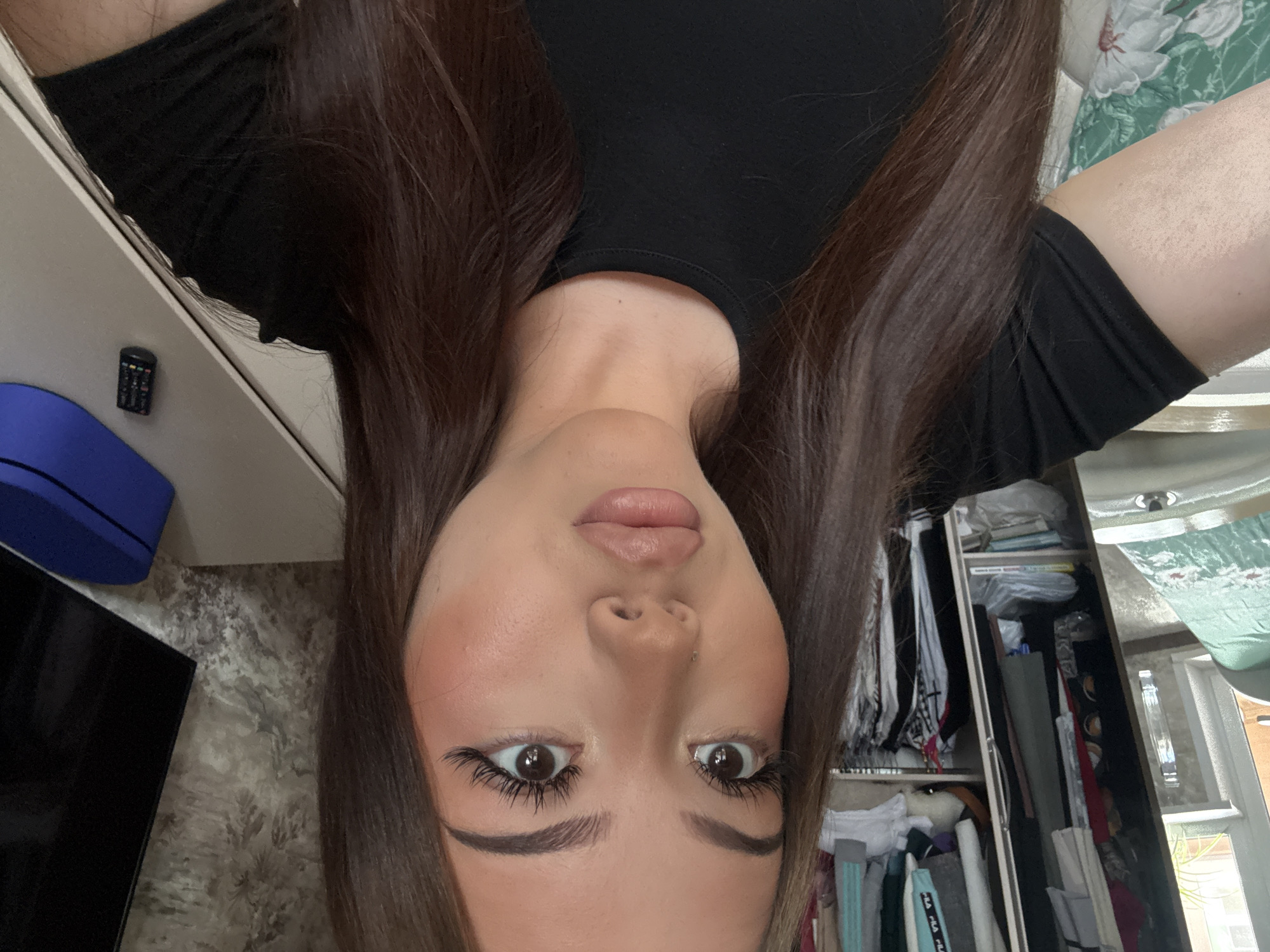 The original image is an upside-down, close-up shot of a young woman from a high angle, likely a selfie, taken in an indoor setting. The woman has long, dark brown hair, falling on both sides of her face, and wears a black top. Her face features prominent makeup, including winged eyeliner, defined eyebrows, mascara-enhanced lashes, and nude-toned lipstick. A small piercing is visible above her upper lip. Her eyes are wide open, looking directly towards the camera.
The background, also inverted, reveals details of a room, consistent with a bedroom in Naberezhnye Chelny, Russia. To the viewer's left (which would be the woman's right side if oriented correctly), a light-colored surface holds a small, black remote control with numerous buttons, positioned next to a blue rectangular object. A dark, flat screen, likely a television, is partially visible further left against a textured, grayish wallpaper. To the viewer's right (the woman's left), an open wardrobe is packed with an assortment of clothes and items. Among the garments, a light blue item clearly displays the brand "FILA." Books or papers are stacked on shelves within the wardrobe. A window in the background, behind the wardrobe, indicates natural daylight. A green fabric with a white floral pattern, possibly a bedspread or curtain, is visible in the upper right. The scene depicts a casual, personal moment captured during the day.