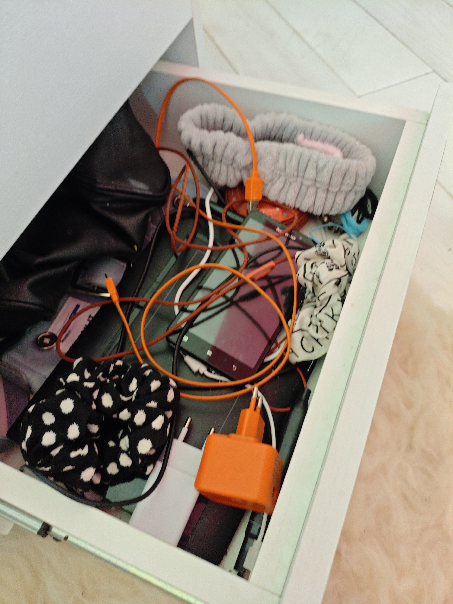 A cluttered open drawer contains various electronic accessories, including charging cables, power adapters, a smartphone, and a scrunchie with a black and white polka dot pattern. A grey, plush headband is also visible, along with a black bag. The drawer itself appears to be part of a white piece of furniture, possibly a desk or dresser, set against a backdrop of light-colored wooden planks. The overall impression is one of disorganization within a personal space, likely a bedroom or living area in a residence located in Трудовское сельское поселение, Ukraine.