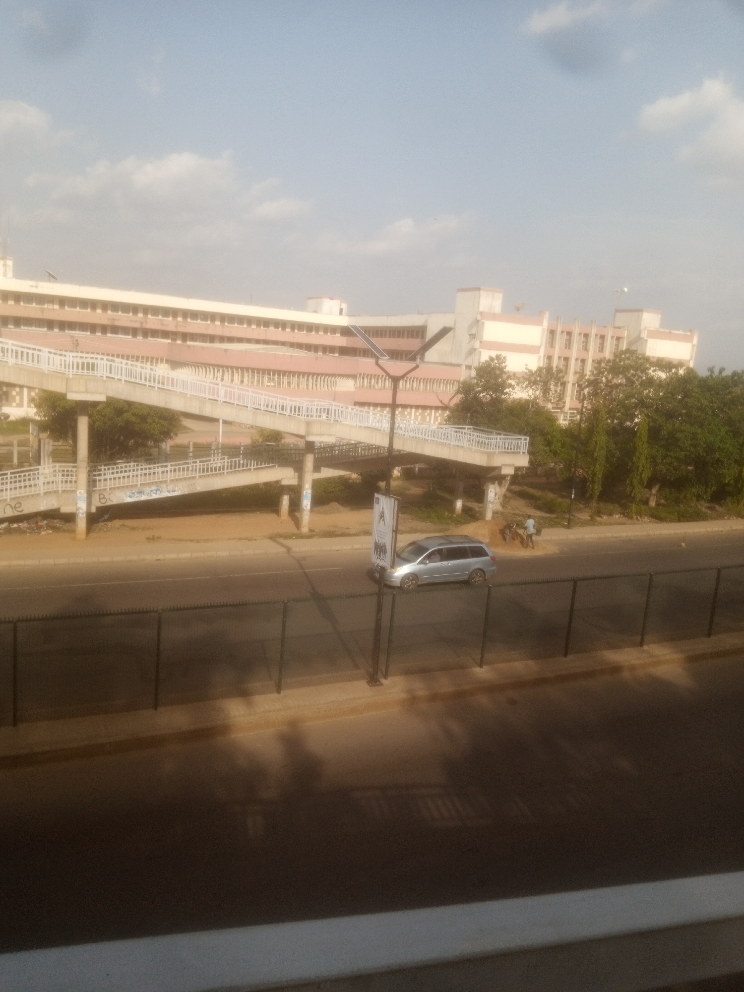 The image shows a pedestrian overpass spanning across a road in Unguwan Dosa, Nigeria. A silver minivan is driving on the road beneath the overpass. In the background, a large, multi-story building with light-colored walls and rows of windows is visible. Lush green trees surround the building and the overpass. The sky is partly cloudy, suggesting it is daytime. The scene appears to be a busy urban environment with infrastructure for both pedestrians and vehicles. There is some graffiti on the supports of the overpass. A tall pole with a sign is visible on the left side of the overpass. There are a few people visible near the road, possibly crossing or observing the traffic.