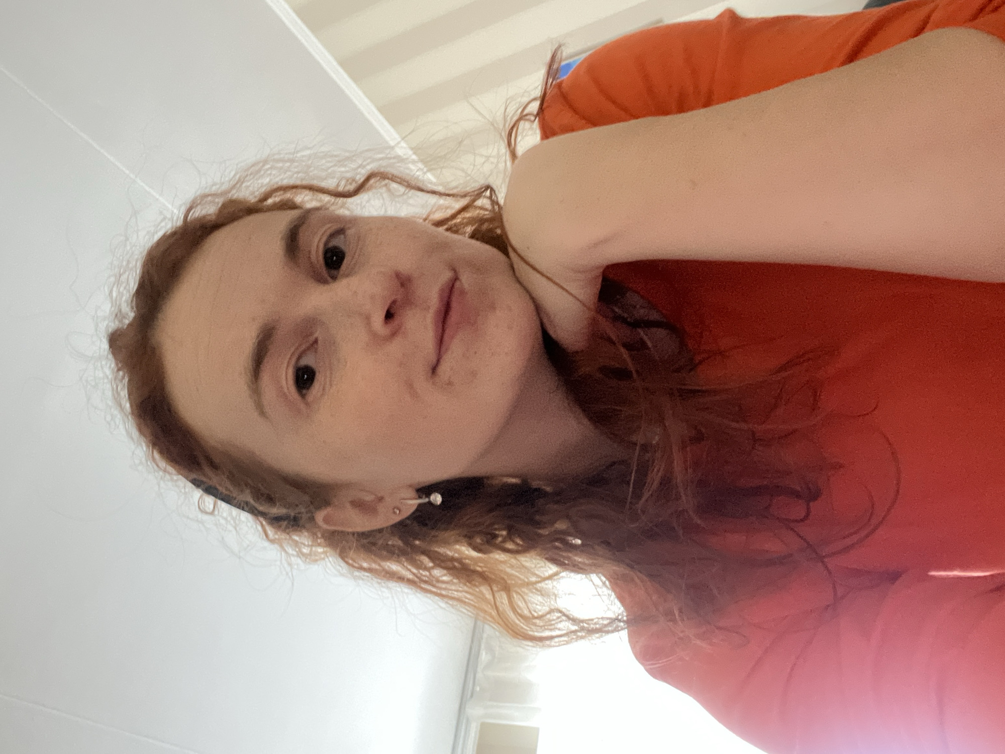 The image is an angled, close-up selfie of a young individual with reddish-brown, wavy to curly hair and fair skin, featuring visible freckles across the nose and cheeks. They are looking directly at the camera with a neutral to slightly soft expression.
The person is wearing a bright orange or reddish-orange t-shirt or top. A small, light-colored earring, possibly a silver stud or pearl, is visible in their left earlobe. A dark band, possibly a hairband or scarf, is partially visible under their hair near the left side of their head. One arm is bent, with the hand resting near their neck or shoulder, suggesting a relaxed pose, possibly while lying down or leaning back.
The background is predominantly a bright, light color, appearing to be an indoor setting. Faint horizontal lines are visible in the upper part of the background, which could be architectural details like ceiling panels or wall textures, or possibly window blinds. The lighting is bright and even. There is no visible text in the image. This scene takes place in Chaikovsky, Russia.