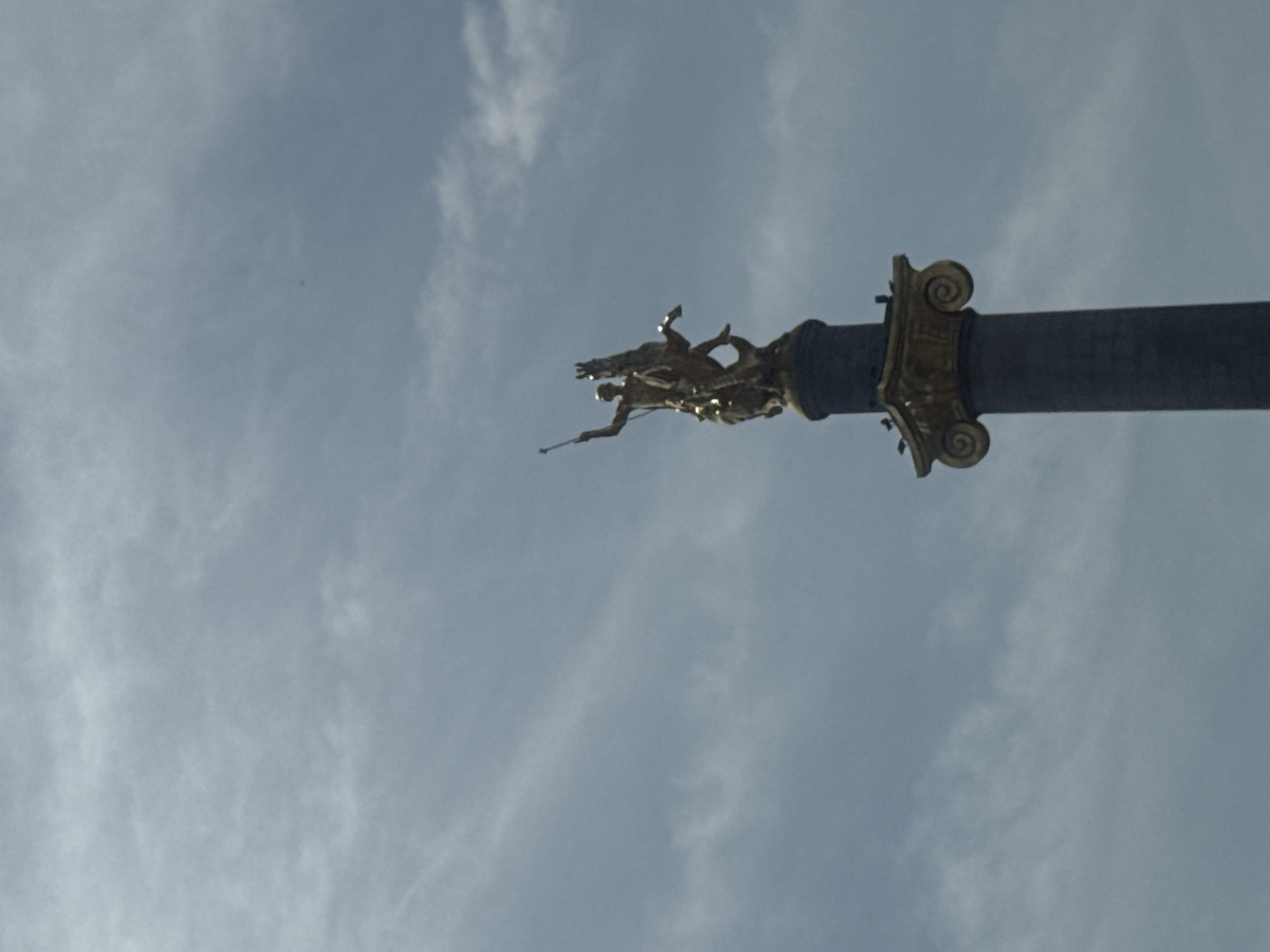The image depicts the St. George Statue atop the Freedom Monument in Tbilisi, Georgia, seen from below against a backdrop of a cloudy sky. The statue, rendered in gold, features St. George slaying a dragon. The monument's column is dark and cylindrical with a decorative, Corinthian-style capital in gold.