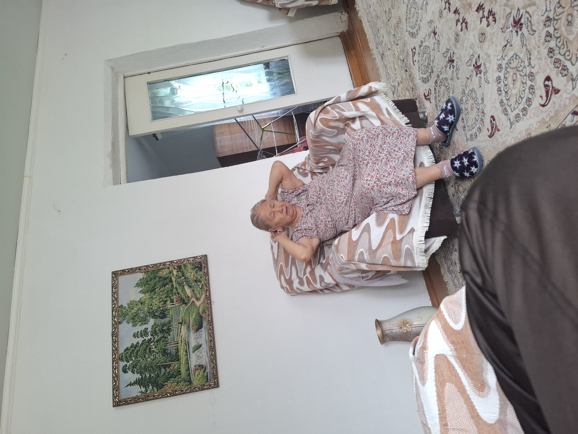 An elderly woman sits in a floral-patterned armchair in a room. She is wearing a floral dress and slippers, and her hands are behind her head. A patterned blanket is draped over the armchair. To her left, on the floor, is a decorative vase. On the wall behind her is a framed landscape painting of a forest and waterfall. To the right of the armchair is a door with a glass panel, and beyond the door, a wooden cabinet is visible. The room has a patterned rug on the floor and white walls. The image was taken indoors.