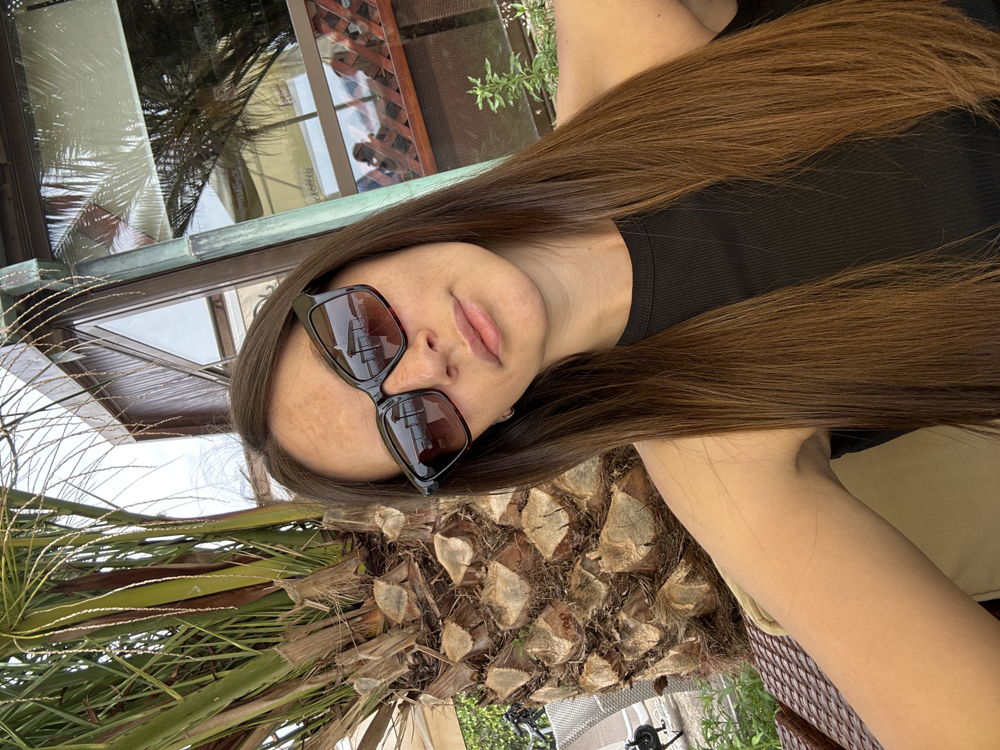 A woman with long brown hair is wearing sunglasses and a black shirt. She is outdoors, near a palm tree. The image appears to be taken in Budva, Montenegro, suggested by the vegetation and architectural details visible in the background.