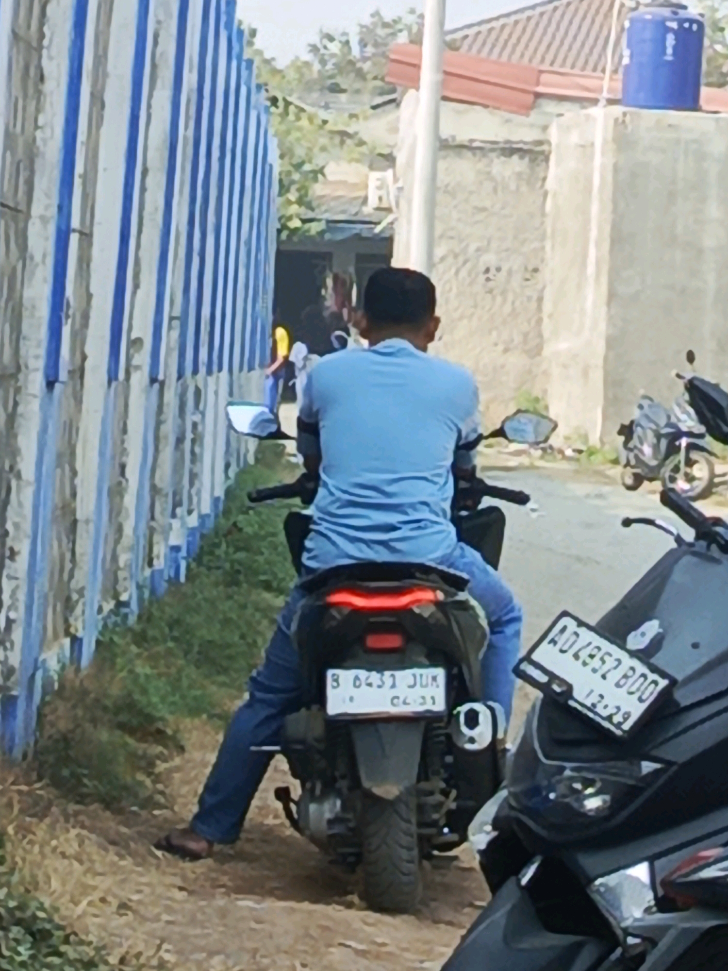 The image shows two motorcycles parked on a dirt path next to a fence with blue and white stripes, located in Jati Mulya, Indonesia. The motorcycle in the foreground, on the right, has a license plate reading "AD 4352 BOO 12-29". A person wearing a blue shirt and blue jeans is seated on the motorcycle in the background, which has a license plate "B 6431 JUK 04.21". The background includes buildings with tiled roofs and trees. The scene appears to be outdoors on a sunny day.