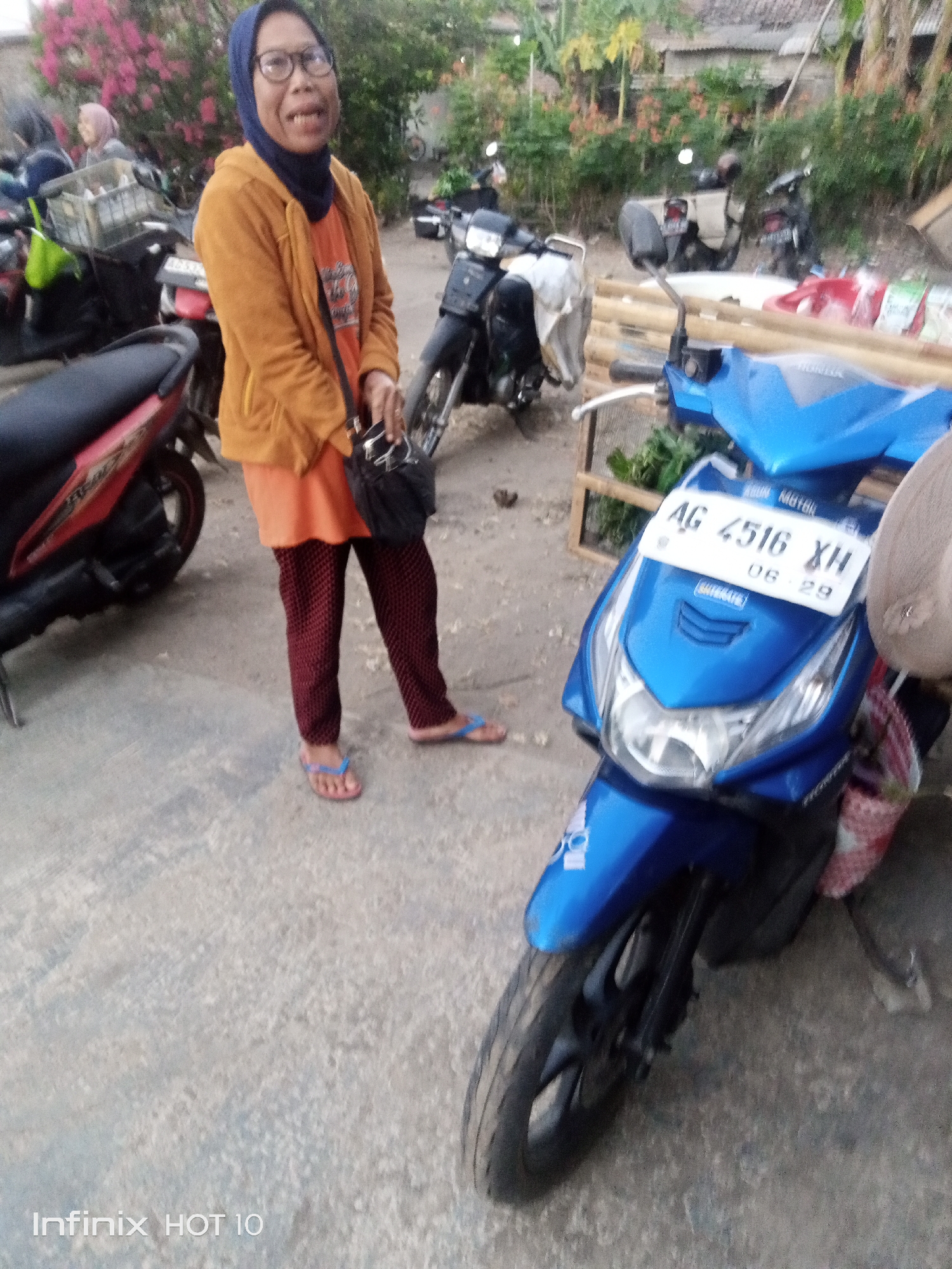 In the city of Sono Ageng, Indonesia, an Indonesian woman wearing glasses, a blue hijab, an orange shirt, and patterned red pants stands beside several motorcycles. She is looking towards the camera with an open mouth, appearing to speak or laugh. She is holding a black bag with a strap over her shoulder. A red motorcycle is visible to her left, and a bright blue scooter with a license plate reading "AG 4516 XH" is in the foreground. The background shows other motorcycles, some vegetation, and buildings with corrugated metal roofs, indicating a residential or semi-urban area. The ground is paved with concrete.