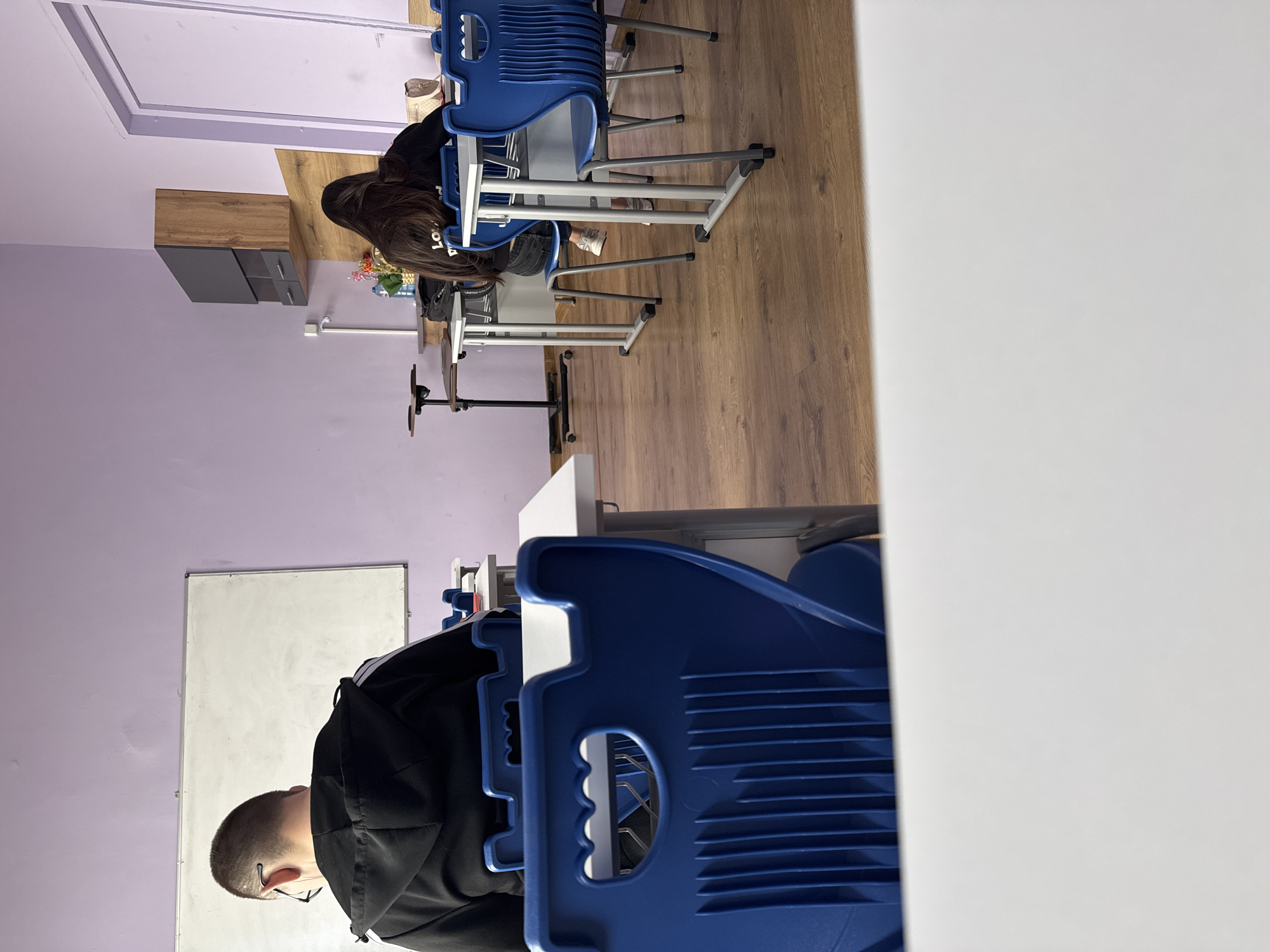This image depicts an indoor scene, likely a classroom or study area in Buhovo, Bulgaria. The walls are painted a light lavender color, and the floor is made of wooden planks.
Two individuals are visible, seated at desks. In the foreground, positioned to the left (after mentally re-orienting the image), a person with short, buzzed hair and glasses is seated at a white-topped desk, wearing a black hoodie. Their back is mostly towards the viewer, facing a large white board mounted on the wall. Further into the room, to the right, another person with long, dark hair is seated at a similar white-topped desk. They are wearing a black long-sleeved top with partial text "LOV" and possibly "ED" visible on the back, suggesting "LOVED" or similar. A bag with text that appears to be "UNITED CO" (likely part of "UNITED COLORS OF BENETTON") is visible near their feet.
Both individuals are seated on blue chairs with metal frames, typical of classroom furniture. In the background, there is a wooden cabinet or credenza with dark gray drawers or doors, on which a small plant in a pot and a water bottle are placed. A white power outlet is visible on the wall near the cabinet. The overall setting suggests a quiet learning or working environment. No specific time of day is discernible from the indoor lighting.