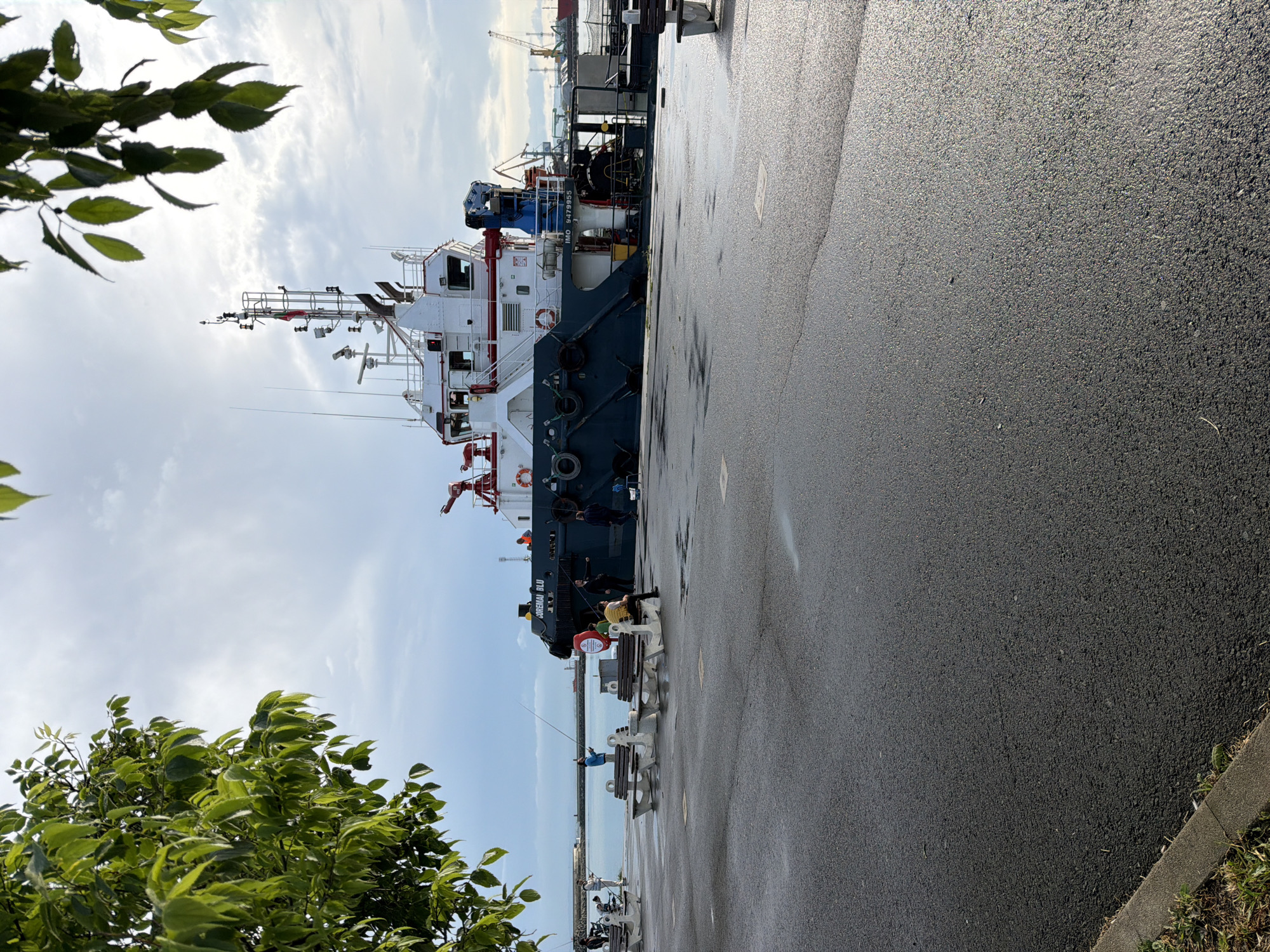The image shows a tugboat docked at a port in Burgas, Bulgaria. The tugboat is primarily white and blue, with visible safety equipment like life rings and red fire hoses. The name "CHREMAI BLU" is visible on the hull. The IMO number 9479955 is also visible. Alongside the dock, people are fishing from benches. The ground is paved with asphalt. In the background, other port infrastructure, including cranes, is visible. Green foliage is visible on the left side of the frame. The sky is cloudy.