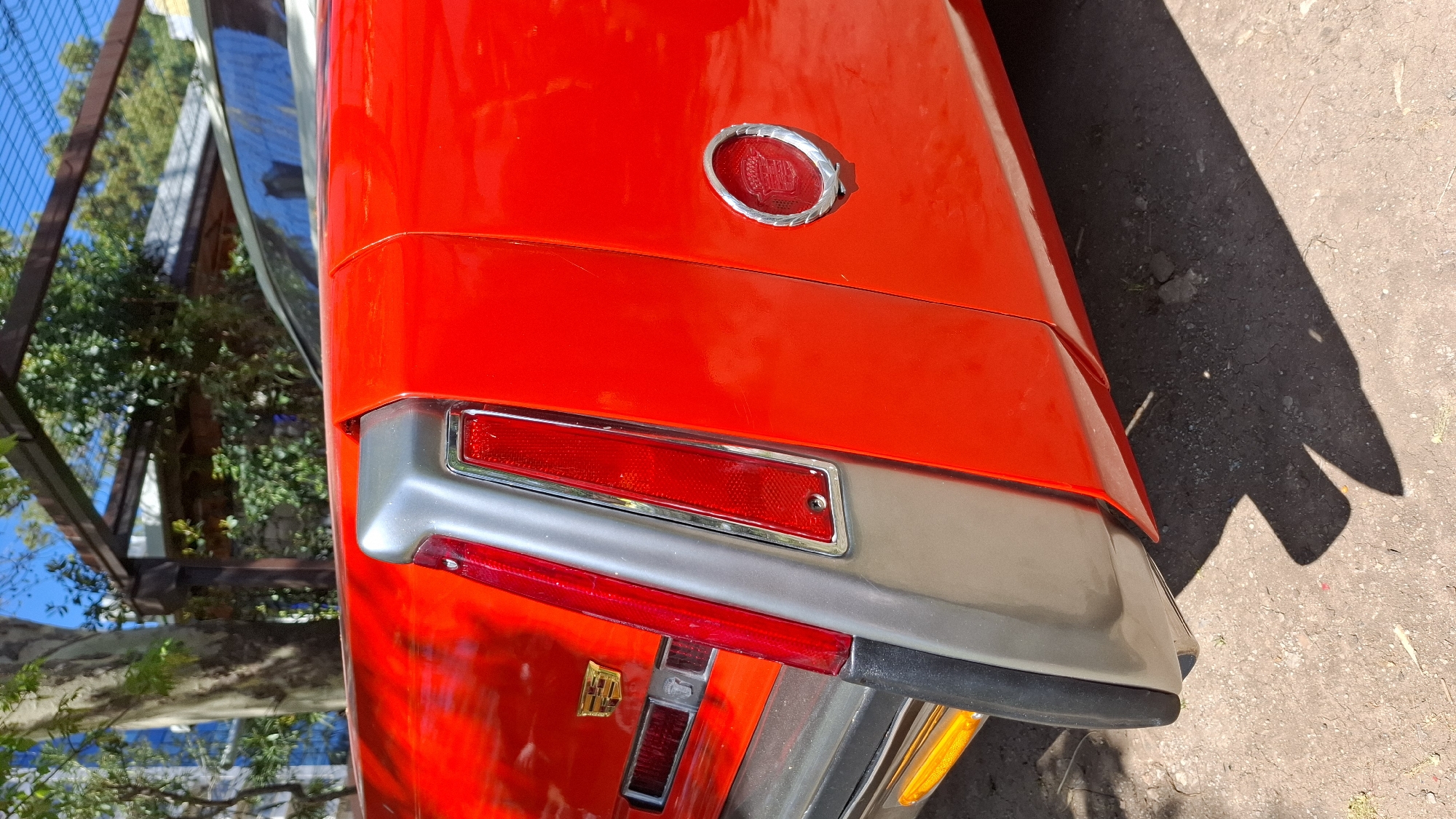 A close-up shows the rear of a red vintage Cadillac automobile, parked outdoors on pavement. The image captures details like the Cadillac emblem, tail lights, and chrome accents. The background shows a tree, plants, a metal fence, and glimpses of a blue structure. The location is not explicitly evident from the visual content, and there is no way to confirm it is in the city of Budva, Montenegro.