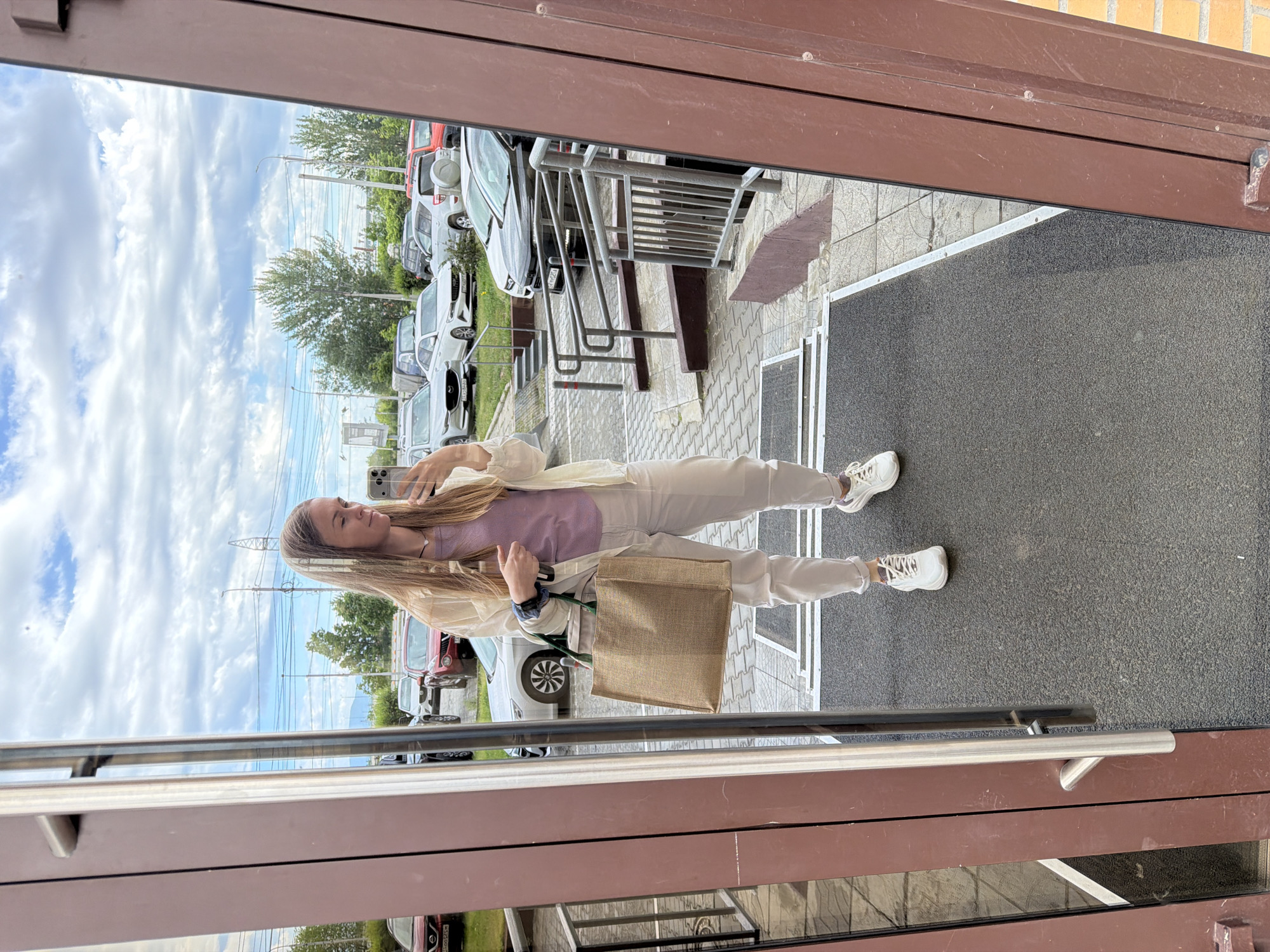 A young woman with long blonde hair, wearing a light beige open jacket, a lavender t-shirt, and light-colored pants, stands in front of a large reflective glass door, taking a selfie with her phone. She is holding a large, rectangular, light brown woven tote bag in her left arm. She is wearing white athletic shoes. The reflection in the glass shows a parking lot with several cars, including a white sedan, a dark SUV, and a red SUV. Overhead, a partly cloudy sky is visible, with telephone wires and a lamppost in the background. The area immediately in front of the door appears to be an entrance with a ramp and grey non-slip mats. The building's exterior is a dark reddish-brown color. This scene is consistent with an urban or suburban setting in Khabarovsk, Russia.