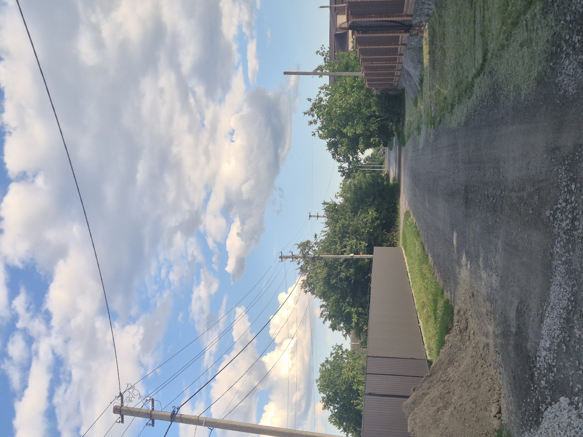 The image depicts a rural road in Sozonivka, Ukraine, under a sky filled with scattered white clouds against a blue background. The road, surfaced with gravel and dirt, stretches into the distance and is lined on both sides by fences and greenery.
On the left, a tall, grey utility pole stands prominently with multiple wires extending from it. Adjacent to the pole is a grey corrugated metal fence. A pile of light brown soil sits on the grass next to the fence. The area is characterized by a mixture of short green grass and some scattered gravel.
On the right side of the road, a fence made of brown wooden planks, set against a stone foundation, is visible. Beyond the fence, more grass and some trees can be seen.
The road is flanked by lush green trees, some of which are tall and mature, creating a natural canopy effect further down the lane. The presence of utility poles and wires suggests that this is a populated area, likely a village or a residential outskirts. In the distance, a small vehicle can be seen on the road. The overall scene conveys a sense of a quiet, possibly developing, residential area in a rural setting.
