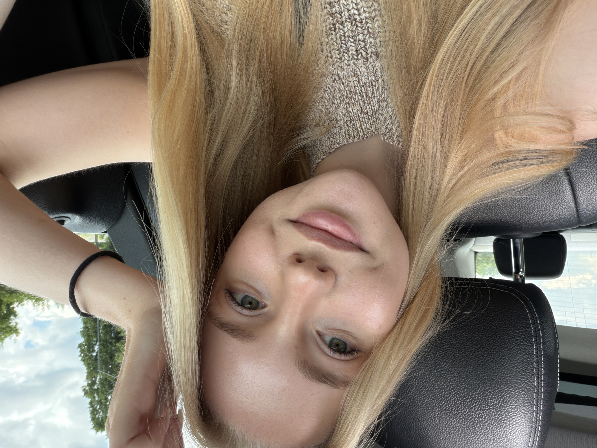 A young Caucasian woman with long, straight, light blonde hair, fair skin, light green eyes, and natural pink lips poses for a selfie inside a vehicle. She has a slight smile and subtle freckles on her face. She is wearing a light brown and beige heathered knit sweater. Her left arm is raised, with her hand positioned near the top of her head, mostly out of frame, while a black hair tie is visible on her right wrist. The interior of the vehicle features a black leather headrest with white stitching to her left, and black interior panels. Through the windows in the background, green trees and a cloudy sky are visible, indicating an outdoor, possibly suburban or rural, setting.