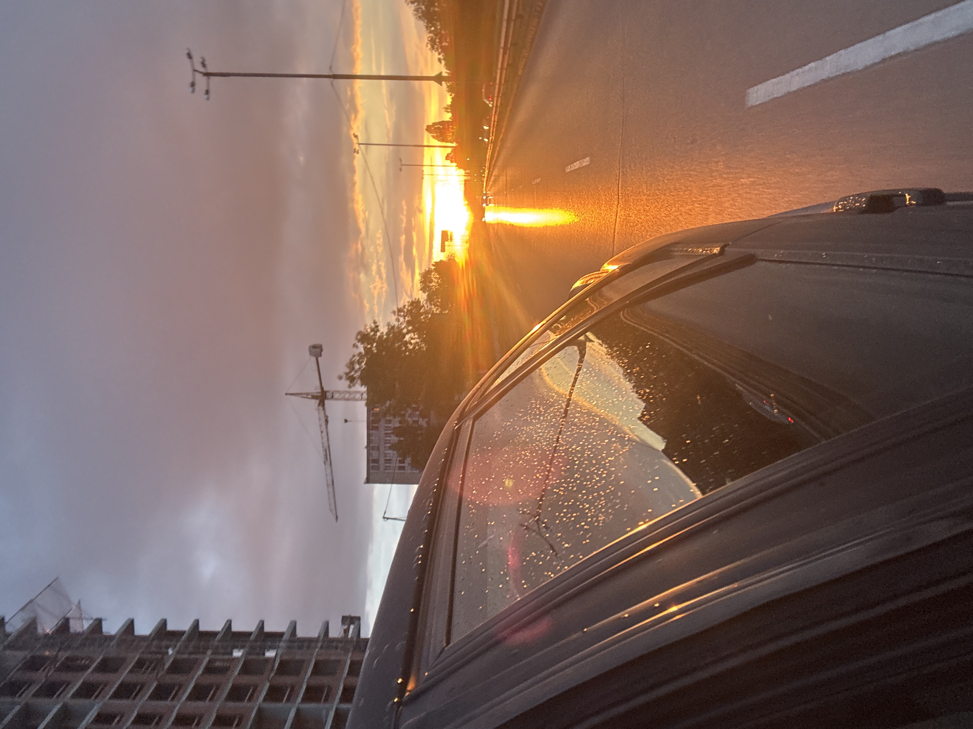 The image is taken from inside a car, likely in motion, with a view through the passenger window. Raindrops are visible on the window. The scene appears to be near a construction site, as a partially built structure and a crane are visible. Across the street, a highway and some commercial buildings are also visible. The sunset is reflecting off the road and the car, creating a golden hue. There are other cars driving in the opposite direction. The overall environment suggests an urban setting during sunset or early evening, with overcast conditions. Based on the architectural style and infrastructure, the location is likely in Sofia, Bulgaria.