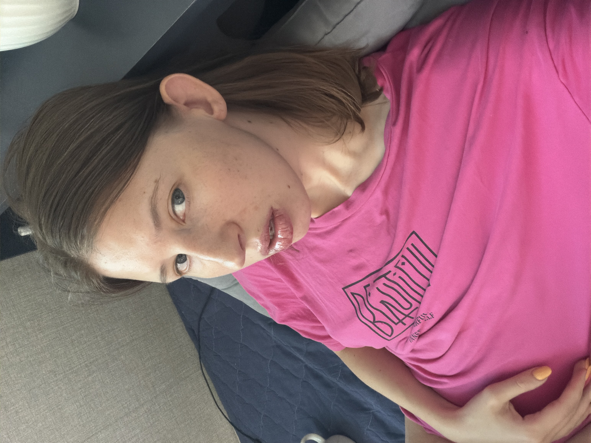 A woman with brown hair and blue eyes, wearing a bright pink t-shirt with a black graphic logo, is lying down indoors. The background shows a textured gray surface and a dark blue quilted fabric. Part of a white object, possibly a mug, is visible on the left. The woman's skin has freckles, and her lips are glossy. Her right hand, with orange nail polish, is resting on her abdomen. The scene appears to be in a residential setting. The image does not contain any discernible information related to Dongducheon-si, South Korea.