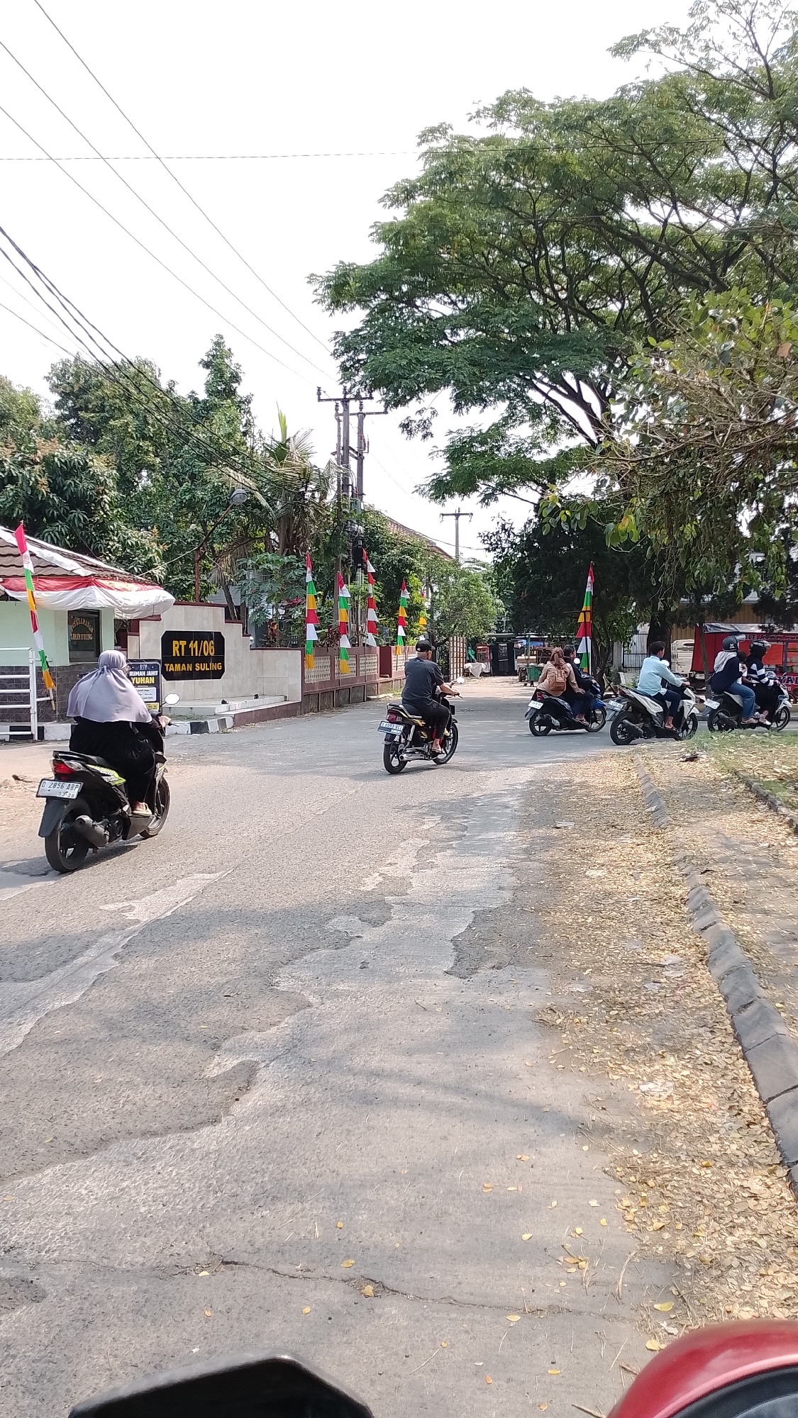 The image depicts a street scene in Bojongsoang, Indonesia, near the entrance to a building identified as "RT 11/06 TAMAN SULING". The road surface is asphalt, showing signs of wear and tear with patches and cracks, and is lined with a concrete curb on the right. Fallen leaves are scattered along the roadside. Several motorcycles are present, with some individuals riding them and others as passengers.
In the foreground left, a person wearing a grey hijab and dark clothing rides a black scooter, license plate visible as "G 2856 ABY". To the right of this scooter, further down the road, a person on a yellow and black motorcycle, license plate visible as "E 4593 BF", is riding away from the camera. In the mid-ground, several other motorcycles are visible. One motorcycle carries two people, with the passenger in front appearing to be a woman with brown hair. Another motorcycle has two individuals, one in a light blue shirt and the other in darker clothing. Further to the right, two more motorcycles are partially visible, one with at least two riders.
The building to the left has a sign that reads "RT 11/06 TAMAN SULING". The building is adorned with Indonesian flags, consisting of red and white stripes, and also with flags featuring red, yellow, and green stripes. Trees, including a large one with dense green foliage on the right, and power lines with utility poles are also part of the environment. The sky is overcast.