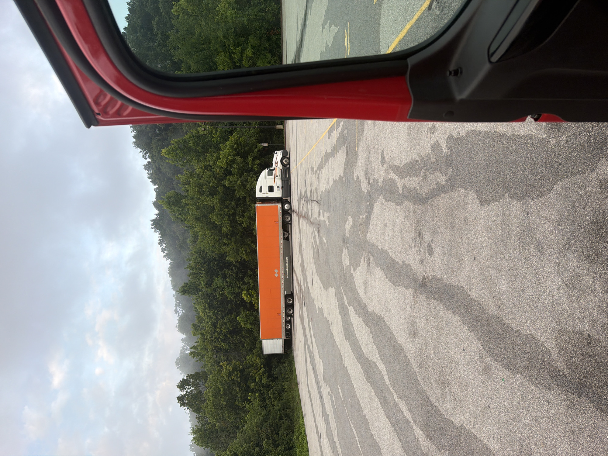 A red truck cab door is open on the right side of the frame, obscuring the foreground. In the middle ground, a white semi-truck with an orange trailer is parked in a lot. The trailer has "schneiderjobs.com" printed on its side. The lot is paved and appears wet, with dark, elongated streaks across the surface. In the background, a dense line of green trees covers a hillside under a cloudy sky. The scene appears to be in a rural or semi-rural area. The exact location in Wildwood, United States is not discernible from the image alone.
