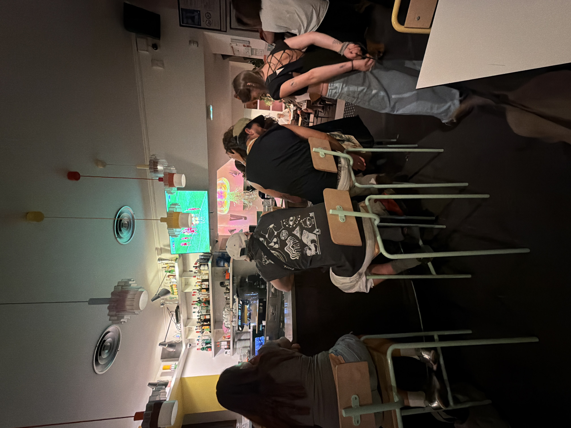 This is a photograph taken indoors, likely in a bar or restaurant, with a modern, possibly industrial aesthetic. The lighting is dim, with spotlights and overhead fixtures contributing to the atmosphere. In the foreground, there are several people seated at a bar, facing away from the camera. The bar area is stocked with bottles and glassware, suggesting it's a functional establishment. A television screen is visible in the background, displaying a sports game, indicating entertainment is available. The people are dressed casually, and their interactions are not clearly discernible due to their postures and the angle of the shot. The overall environment appears to be a social gathering space, likely within a city. There is no specific visual information in the image that would allow for the identification of the city of Lyon, France.