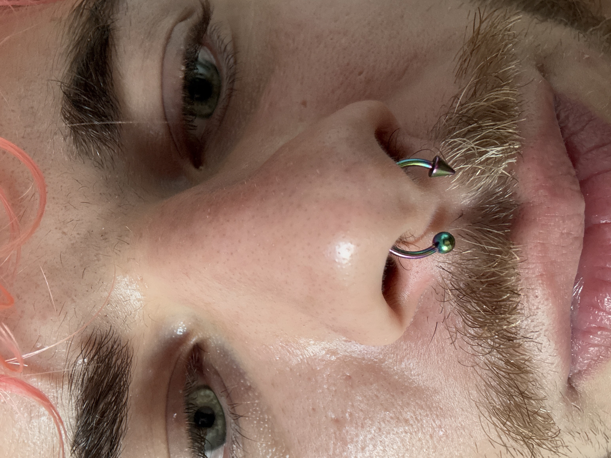 A close-up image, taken at an unusual angle, shows the upper portion of a human face. The face is pale with sparse blond facial hair growing from the upper lip. The left nostril is pierced with a curved, metallic ring with a conical end. The upper right portion of the image includes an eye with dark eyelashes. The upper left portion of the image shows a partially visible eye with dark eyelashes and light pink hair. The image does not provide any information about the location of Đà Nẵng, Vietnam.