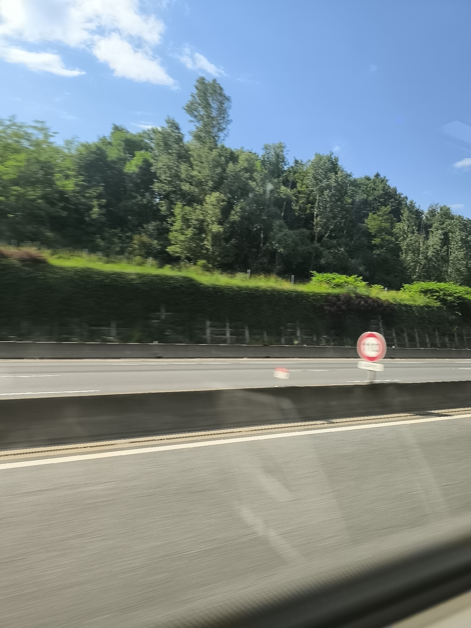 The image shows a view from a moving vehicle, likely on a highway. The foreground is dominated by the blurred asphalt road surface. A concrete barrier separates the road from the greenery on the other side. Beyond the barrier, there's a dense line of trees and shrubs. A circular road sign with a red border and a white center is visible on the right side of the road, possibly indicating a speed limit. The sky above is blue with some scattered clouds. Based on the information provided, it is not possible to locate the exact location in the city of Saint-Chamond, France, from this image.