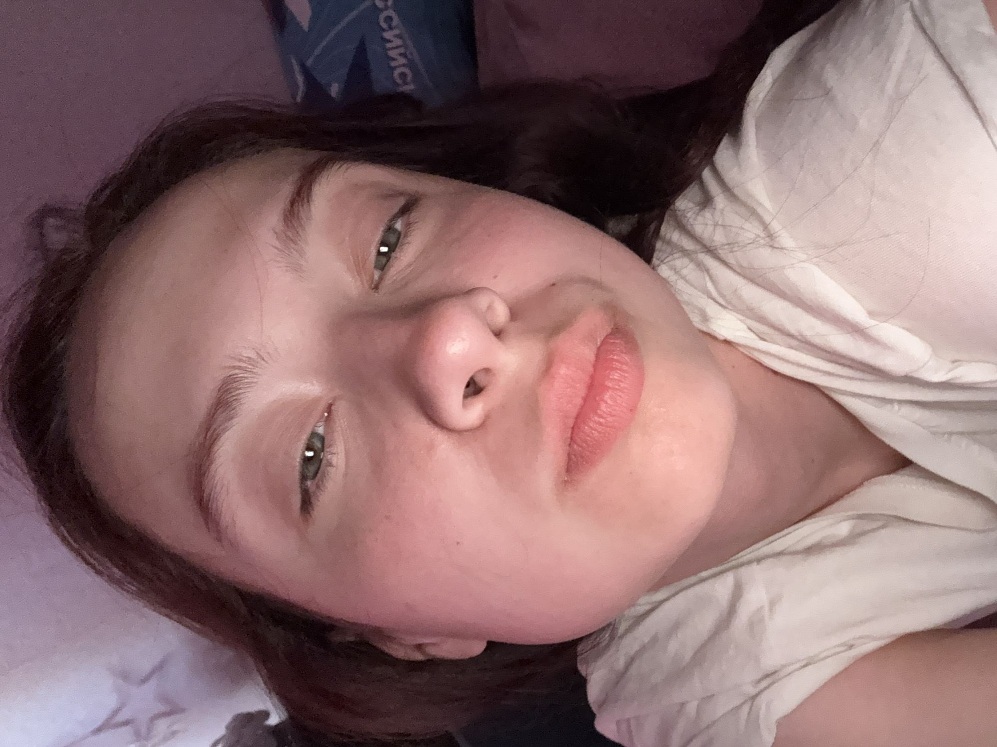 A close-up, slightly angled self-portrait shows a young woman with light skin and dark brown hair. Her eyes are green, and her lips are a natural pink. She is wearing a white t-shirt. The background includes a pillow or bedding with a pattern and a glimpse of a blue object with Cyrillic text. The scene appears to be indoors. The location in the city of Прикамский, Russia is not discernible from the image.