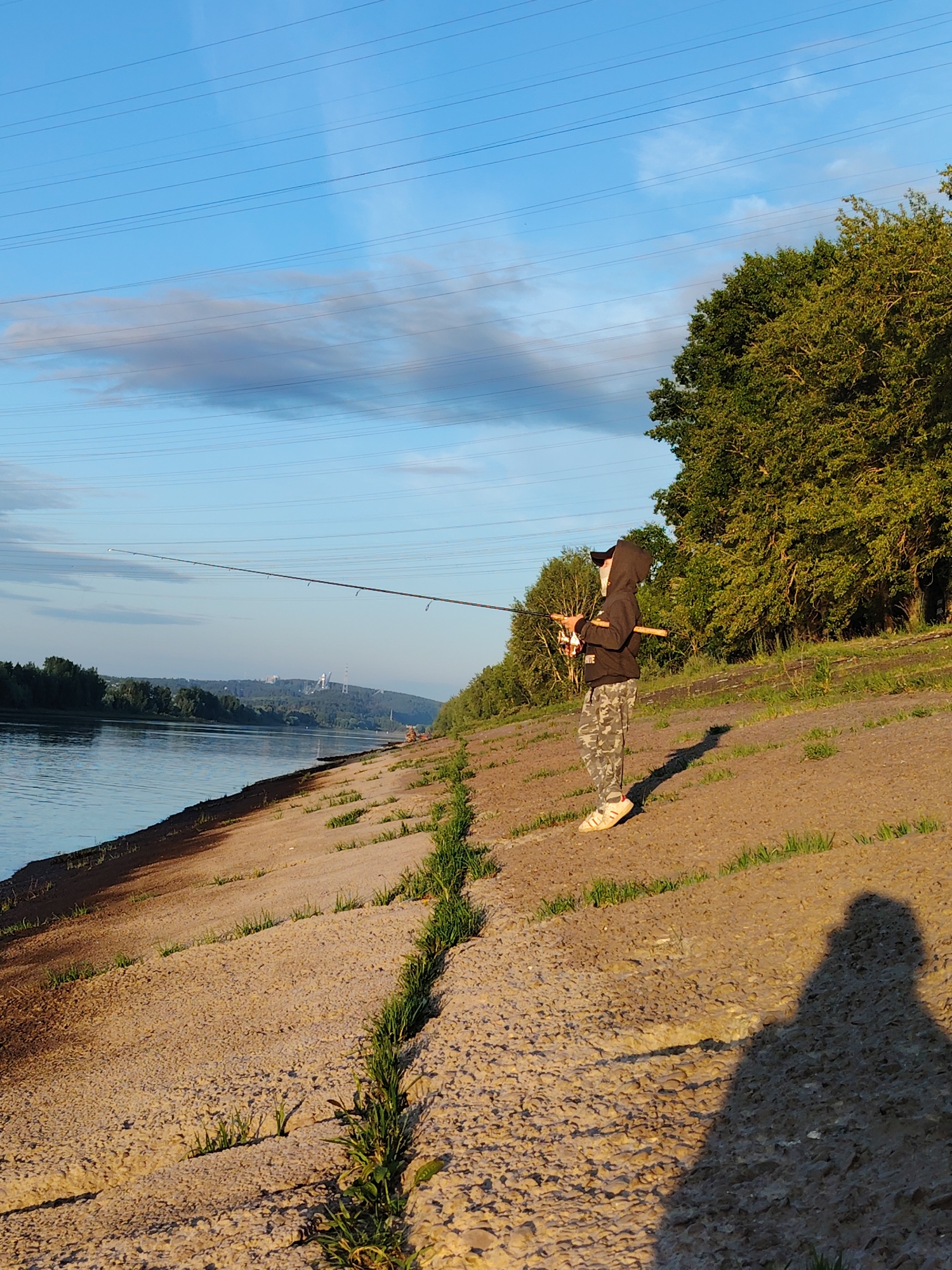 An overhead view shows a lone figure dressed in camouflage pants and a black hooded sweatshirt standing on the concrete bank of a wide, calm river. The person holds a fishing rod, casting a line into the water. The sun casts a long shadow on the bank, indicating late afternoon or early morning. Lush green trees line the opposite bank of the river, and in the distance, industrial structures with tall chimneys are visible. The sky is a clear blue, with scattered wispy clouds and overhead power lines running horizontally across the frame. The scene is set in Chaikovsky, Russia, a city known for its location on the Kama River.