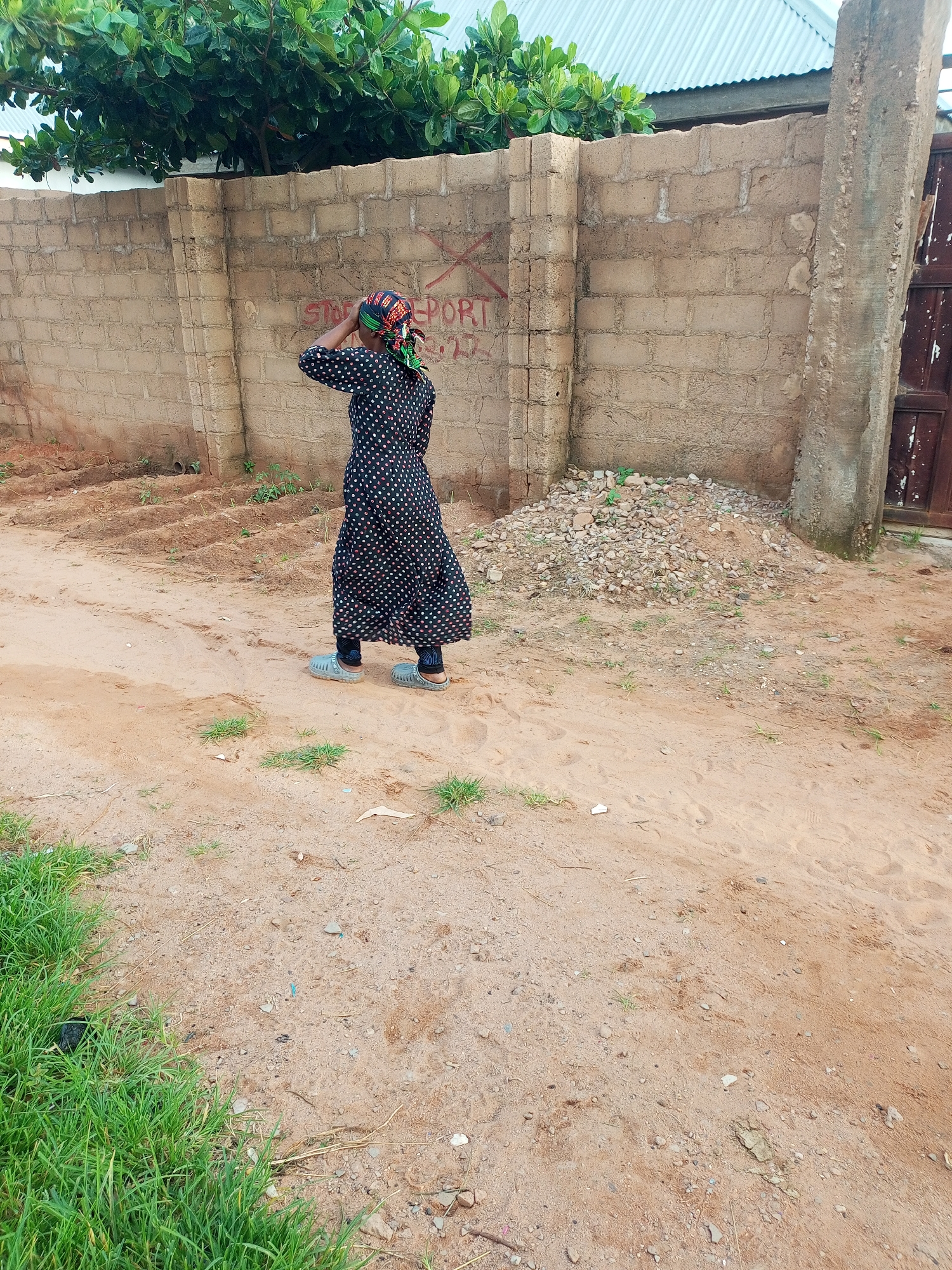 In the city of Tempure, Nigeria, a woman stands with her back to the camera, facing a brick wall. She is wearing a long, black, polka-dotted dress and a colorful headscarf. Her right hand is raised to her head, as if in thought or distress. The wall behind her is constructed of rough, tan bricks and has some red markings on it that appear to be graffiti. To the left of the woman, a patch of green grass and some trees are visible. To the right of the woman, a pile of gravel and a wooden gate are present. The ground in front of the woman is sandy, with some footprints and scattered small stones. The overall scene appears to be an outdoor, semi-urban environment.