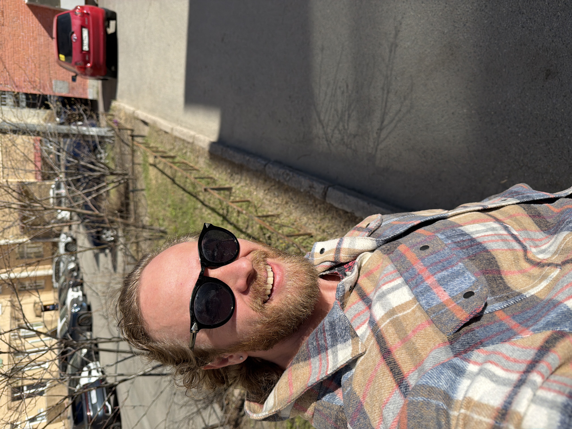 A Caucasian male with a beard and long blond hair is facing the camera, wearing sunglasses and a plaid shirt. The scene appears to be taken from a balcony or elevated position, looking down at a street and parking area in Saint Petersburg, Russia. The photograph captures multiple parked cars, a section of a building with a red roof, and a concrete wall. The lighting indicates a sunny day.