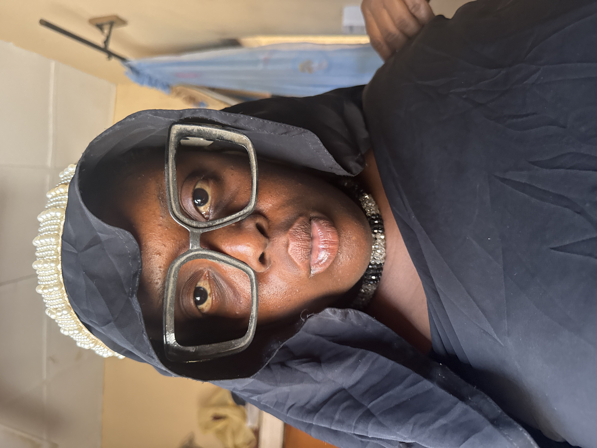 A close-up portrait shot, rotated 90 degrees clockwise, captures an adult individual with dark skin, situated in an indoor setting in Madangala, Nigeria. The person is looking directly into the camera with a neutral to slightly serious expression.
The main subject is wearing large, rectangular-framed glasses with thick, metallic-looking frames. A black head covering, possibly a hijab or shawl, frames the face, and a distinctive white, pearl-like beaded trim is visible along the edge of the head covering on the left side of the frame. Around the neck, a black and silver/clear beaded necklace is visible. The person's lips are slightly parted, revealing a natural sheen.
The background is softly out of focus, showing light-colored walls or ceilings. In the upper part of the image (when correctly oriented), a dark rod or clothesline attached to a wooden block on the wall, and some light blue fabric hanging from it, are visible. The overall lighting suggests an indoor environment. There is no visible text in the image.