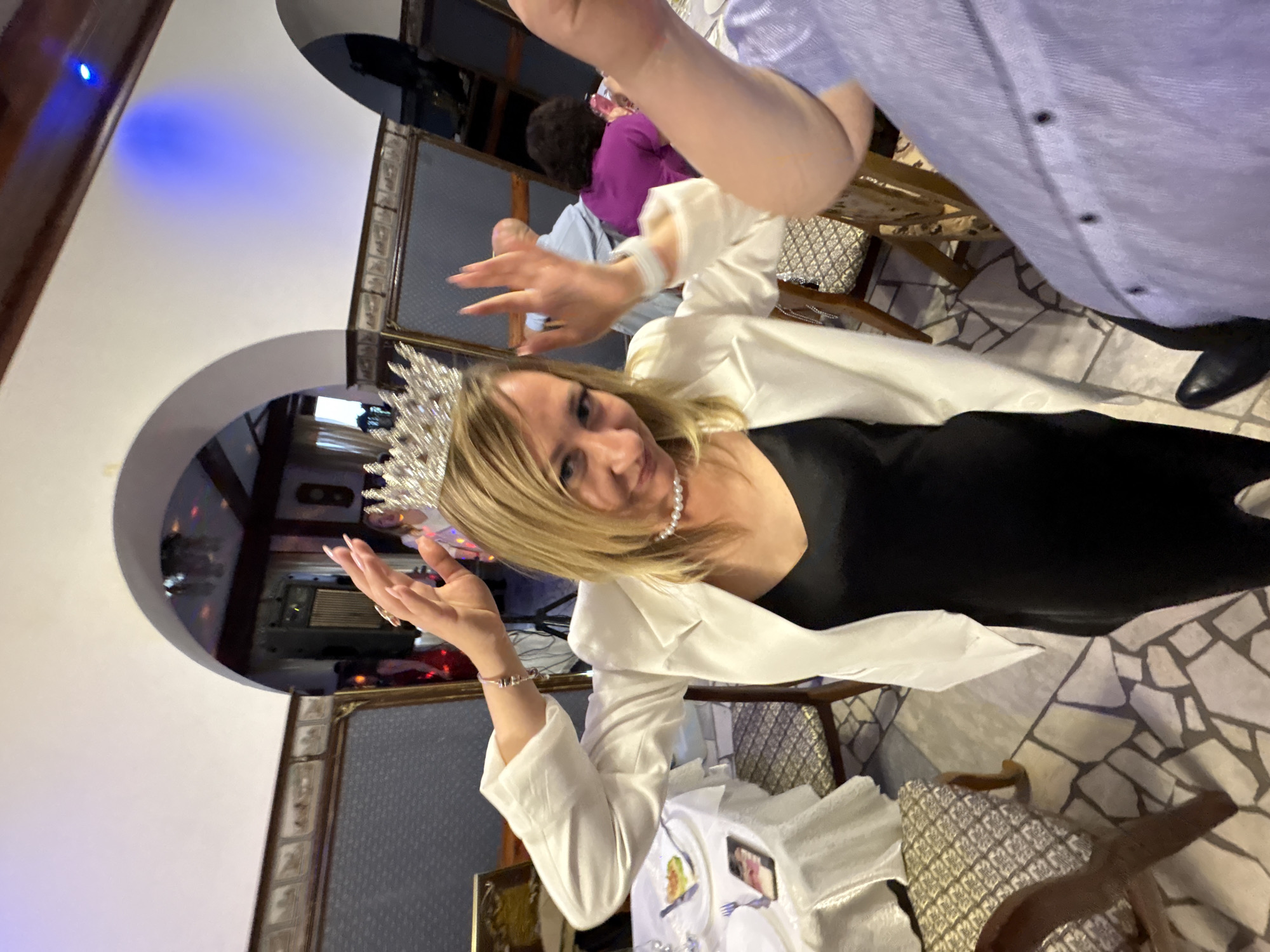 A woman is wearing a tiara and a pearl necklace, and a white blazer over a black dress. She is inside what appears to be a restaurant, possibly in Zavod Mikhailovskii, Russia, based on the user's prompt. The floor is tiled with irregular stone pieces. In the background, there are arches, decorated ceilings with lights, and other patrons at tables. One person's arm and shoulder, wearing a blue shirt, are prominently in the foreground, obscuring part of the woman and the scene. The woman is holding her hands up, near her head, and appears to be posing for the photo. There are tables with white tablecloths set for dining.