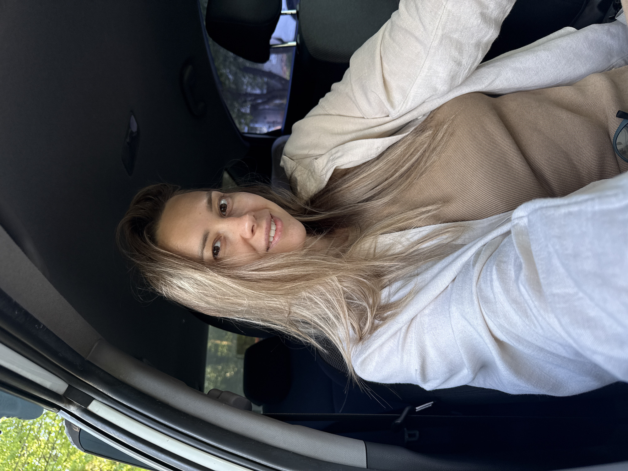 A woman is inside a car, looking at the camera. She is wearing a beige top and a white jacket. The car interior is dark. Outside the car window, trees are visible. The specific location within the city of Chaikovsky, Russia, cannot be determined from the image.