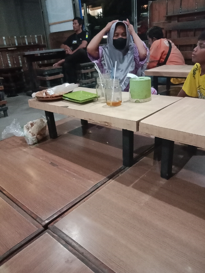 The scene is an indoor or semi-open-air dining establishment, likely a cafe or warung, located in Kebonsari, Indonesia, at night. The flooring consists of light brown wooden panels forming low seating platforms. Low, light-colored wooden tables with dark legs are arranged across these platforms.

In the mid-ground, a woman wearing a light gray hijab, a patterned purple and white top, and a black face mask is seated, her hands raised to adjust her head covering. On the table in front of her are three clear glass jars with white straws, one containing a residual yellowish liquid, two empty brown circular plates, one empty square green plate, and a light green cylindrical tissue dispenser. A clear plastic bag containing a brown pastry item is visible on the floor beside the table's leg.

In the left background, a man in a black short-sleeved shirt with a green emblem is seated at a higher wooden table, looking towards the left. In the right background, a person in an orange long-sleeved shirt is partially visible, facing away. In the right foreground, the head and right shoulder of an individual in a yellow short-sleeved shirt are visible, facing towards the right.