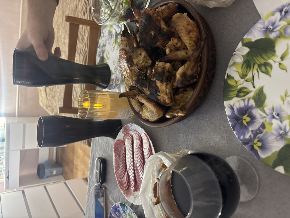 A table is set with food and drinks. A bowl of grilled chicken wings sits beside a floral plate. Slices of salami are displayed on a small dish, next to a glass of red wine. Tall, black vases stand near a lit candle. A hand holds one of the vases. Plates with floral patterns and a salad bowl are also visible. A phone and a small black device are on the table. The scene suggests a meal setting.