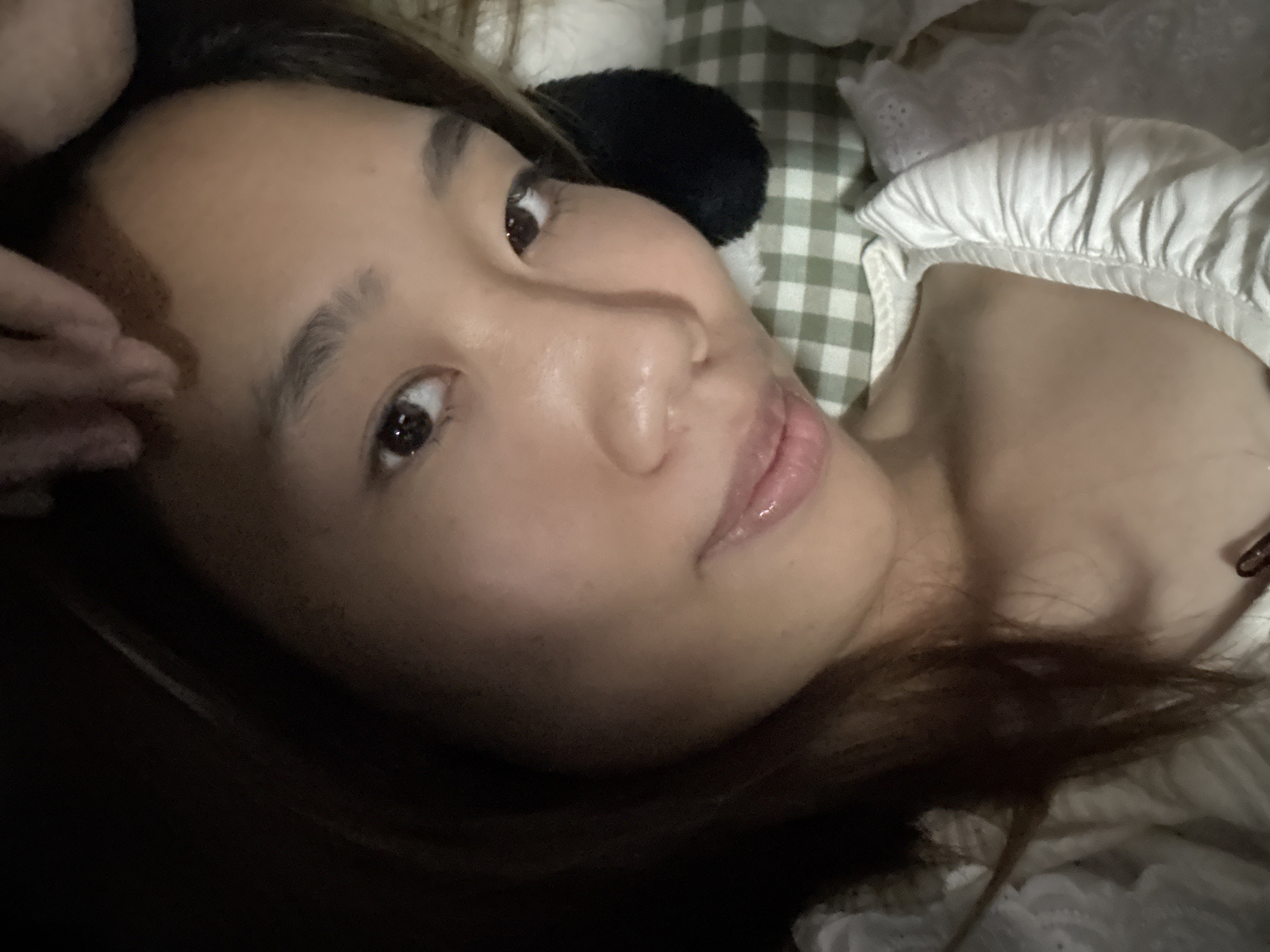 The image shows a close-up, slightly angled view of a woman's face. She has light brown hair and appears to be lying down, possibly on a bed. The background includes a checkered pattern, possibly a blanket or sheet, and some white fabric with ruffles. A dark object, possibly a hair accessory, is near her head. A hand is partially visible at the top left of the frame. The location in the city of Nan'an District, China, is not discernible from the image.