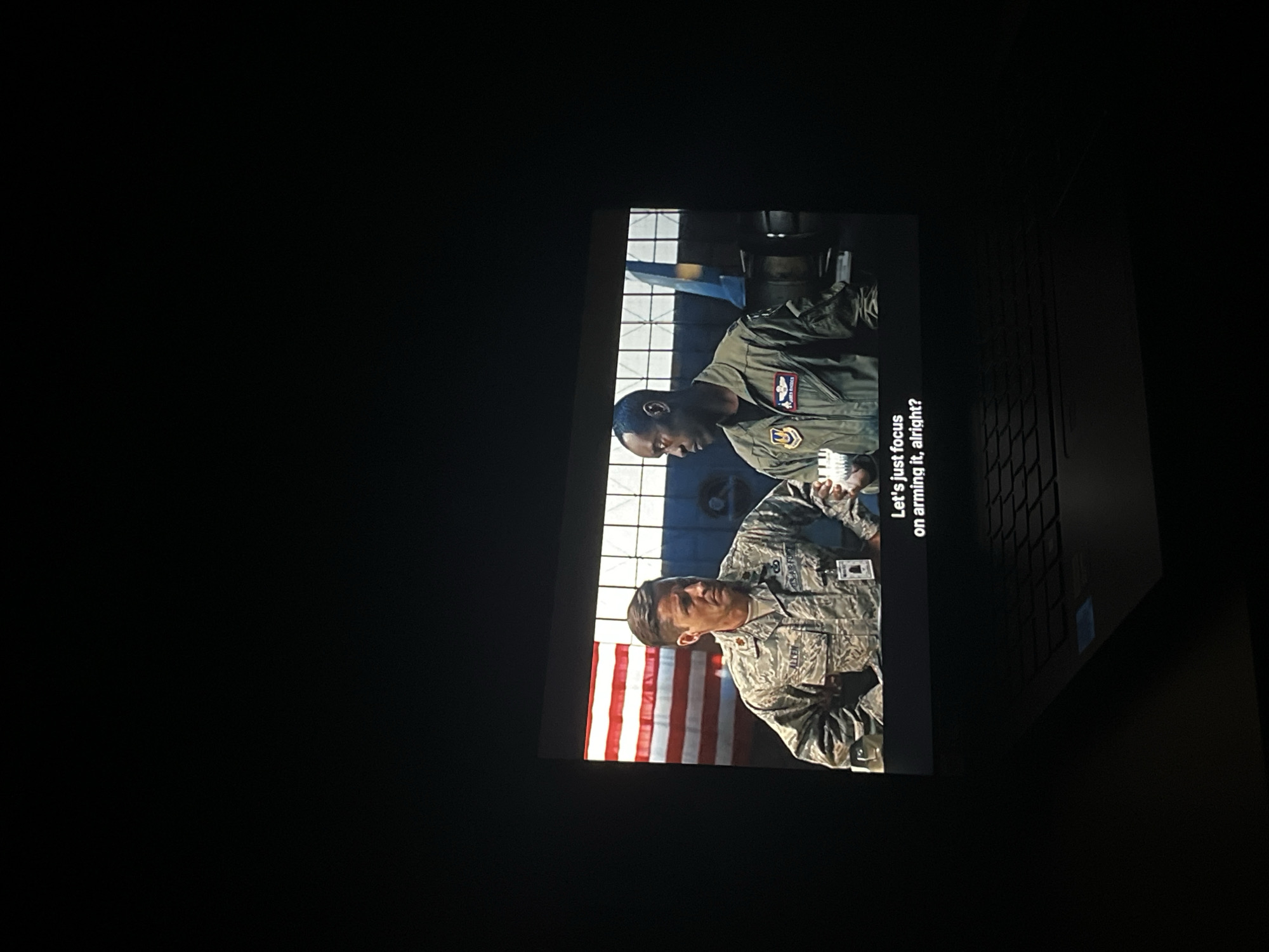 The image shows a screen displaying a scene with two people in military uniforms. The background includes an American flag. A caption reads "Let's just focus on arming it, alright?" The screen appears to be that of a laptop, viewed in a dimly lit environment. There is no information about the city of Sofia, Bulgaria.