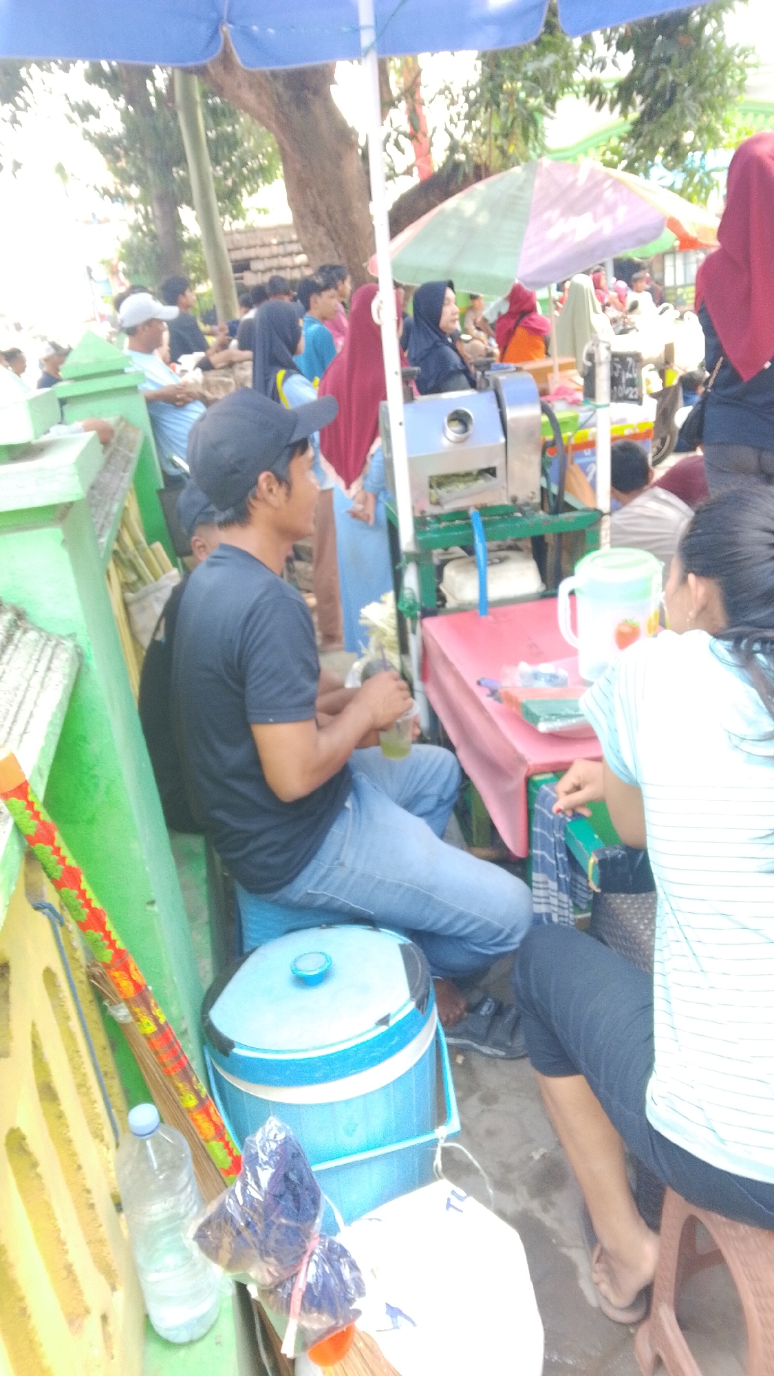 In Mancar, Indonesia, on a sunny day, a man wearing a black t-shirt and blue jeans sits on a blue stool, holding a plastic cup of green liquid. He is at a street-side stall with a sugar cane juicer and a pink table. Several people, including women in hijabs, are visible in the background, some of whom appear to be customers. The stall is under a blue umbrella and has a green fence to its left, adorned with a colorful broom. A large blue cooler is also present. The scene suggests a typical street food vendor operation.