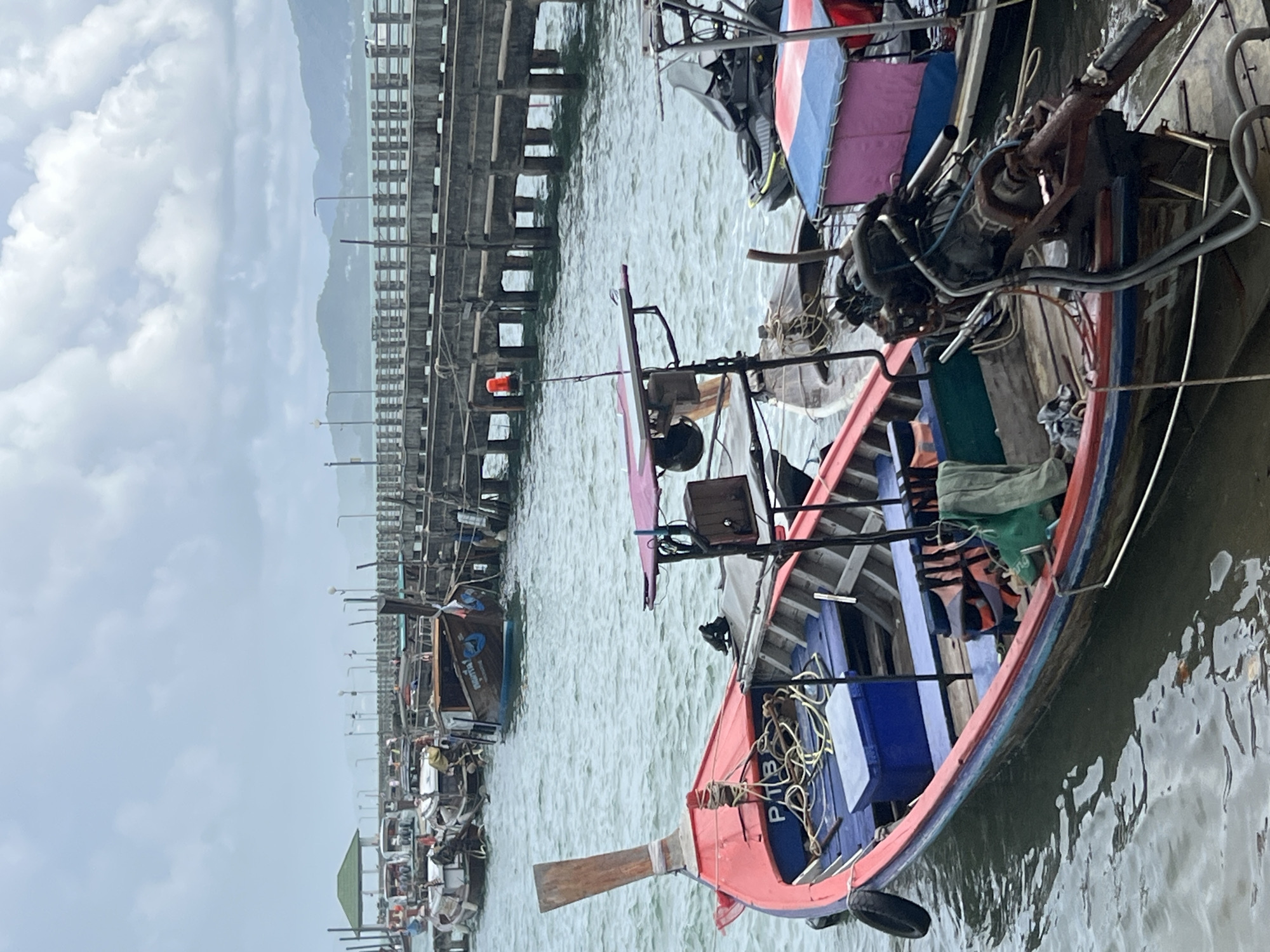 In Pa Khlok, Thailand, a fleet of long-tail boats is docked at a pier under an overcast sky. The boats, painted in vibrant colors like red and blue, are moored alongside a wooden pier that stretches into the distance. The water is calm, with gentle ripples reflecting the muted light. Some boats are equipped with engines, while others appear to be waiting for passengers. The scene is one of quiet activity, with a few boats visible in the background and the pier extending towards a hazy horizon, suggesting a coastal or harbor setting.