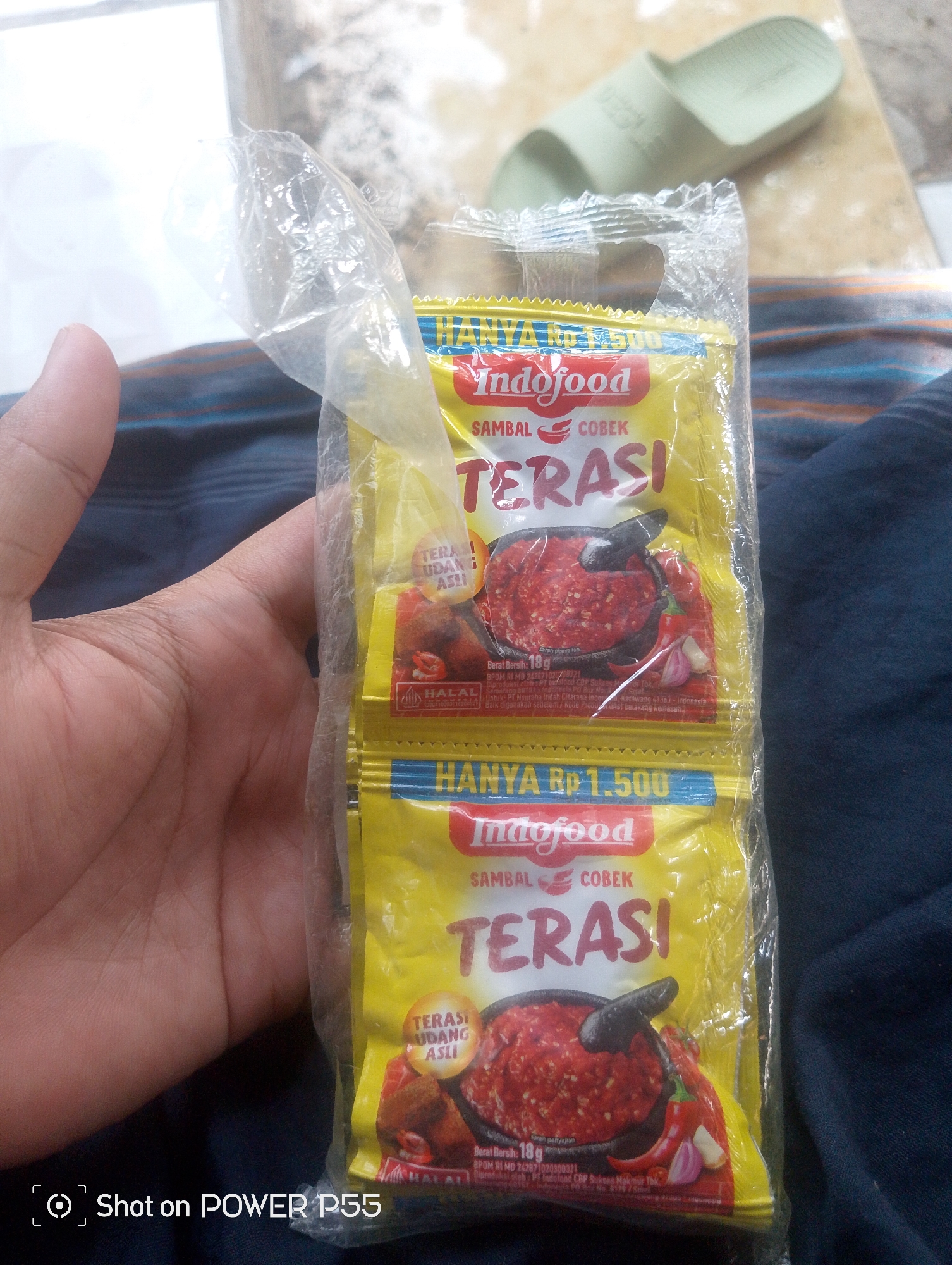 A close-up shot shows a hand holding a clear plastic package containing two sachets of Indofood Sambal Cobek Terasi. The sachets are yellow with red and white text. The upper sachet is partially obscured by the hand. The lower sachet is more visible and features an image of sambal in a mortar, along with chilies and garlic. Below the image, the text "Berat Bersih: 18g" is visible. The words "HANYA Rp 1.500" are printed in blue on the lower part of the package. In the background, out of focus, a pale green sandal is visible, along with a patterned textile. A watermark at the bottom left of the image reads "Shot on POWER P55". The scene is likely indoors, with a focus on the product being held.