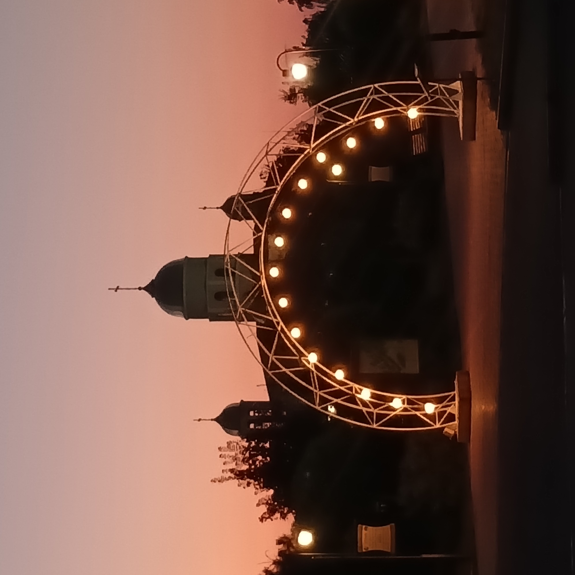 The image captures a twilight scene in Bender, Moldova, featuring a large, illuminated archway decorated with string lights. The archway is a prominent element in the foreground, its metallic structure forming a semi-circle adorned with multiple warm, glowing bulbs. Behind the arch, a silhouette of a church with two domes and a cross atop the central dome is visible against the soft, gradient sky of pink and orange hues characteristic of sunset. Trees partially obscure the church structure, adding depth to the composition. To the left and right of the arch, street lamps with glowing bulbs are also visible, contributing to the overall ambiance of the evening. The ground in front of the arch appears to be a paved surface, reflecting the lights. The scene suggests a peaceful evening setting, possibly in a public square or park.