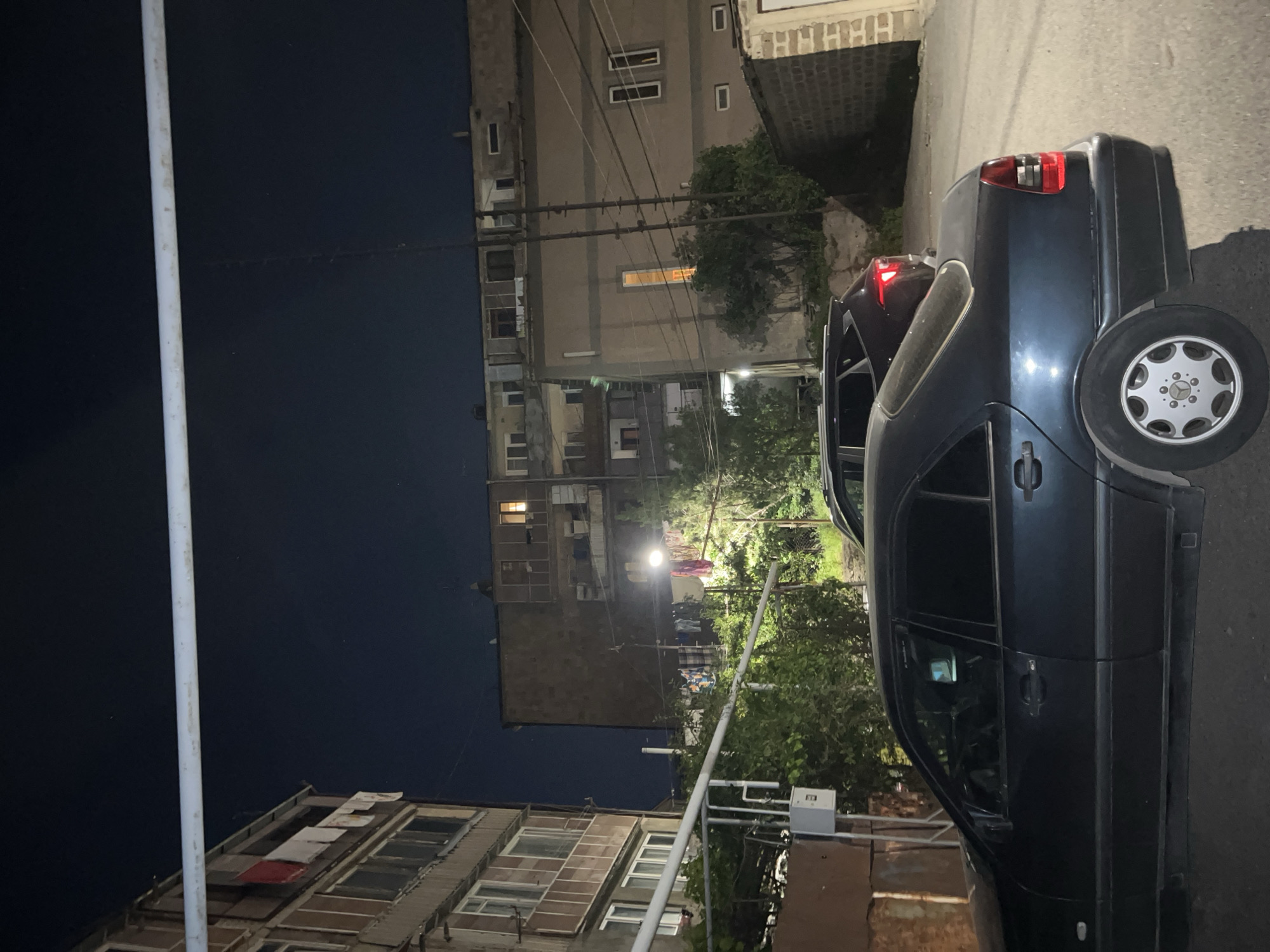 The image presents an urban nighttime scene, likely in Yerevan, Armenia, characterized by a mix of residential buildings and street elements. A dark-colored car dominates the foreground, partially obstructing the view of the street. In the background are multi-story residential buildings with visible windows, some illuminated. Utility poles and wires run between the buildings. In the far background, a street lamp casts a glow, illuminating clothes drying on a clothesline. The overall atmosphere is that of a quiet, possibly low-income neighborhood.