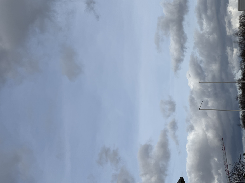 The image depicts an outdoor scene in Oberhausbergen, France, under a partially cloudy sky. The upper two-thirds of the frame show a light blue sky interspersed with various grey, white, and darker cumulus clouds. The lower third of the image primarily features an obscured horizon line with a dense band of dark, leafless trees or shrubs.
In the mid-ground, two tall, slender, light-colored poles stand vertically. The left pole has a horizontal extension projecting to its left from the top. The right pole is a straight vertical column. To the far left, a dark, angled roofline of a building or structure is visible. Below this roofline and above the tree line, the red boom of a construction crane extends diagonally upwards from left to center. A light-colored, flat-topped structure is partially visible on the far right, just above the tree line. No people or active interactions are present in the scene.