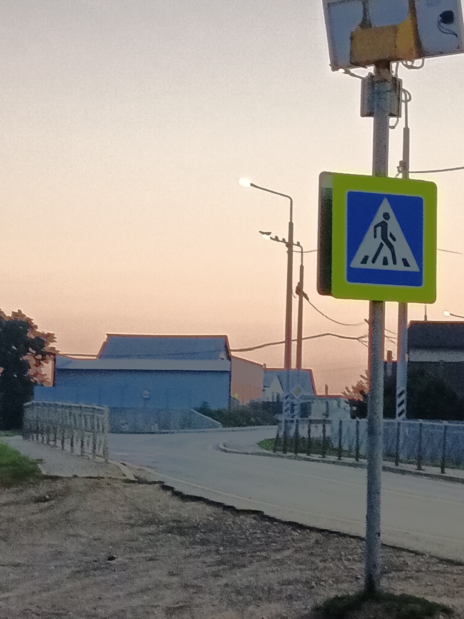 The image shows a pedestrian crossing sign at an intersection in Tatarstanskaya Kargala, Russia. The sign is blue and white with a silhouette of a person walking. It is mounted on a metal pole. In the background, there is a road, buildings, and trees. The sky is overcast, and the sun is setting. There are no people visible in the image.