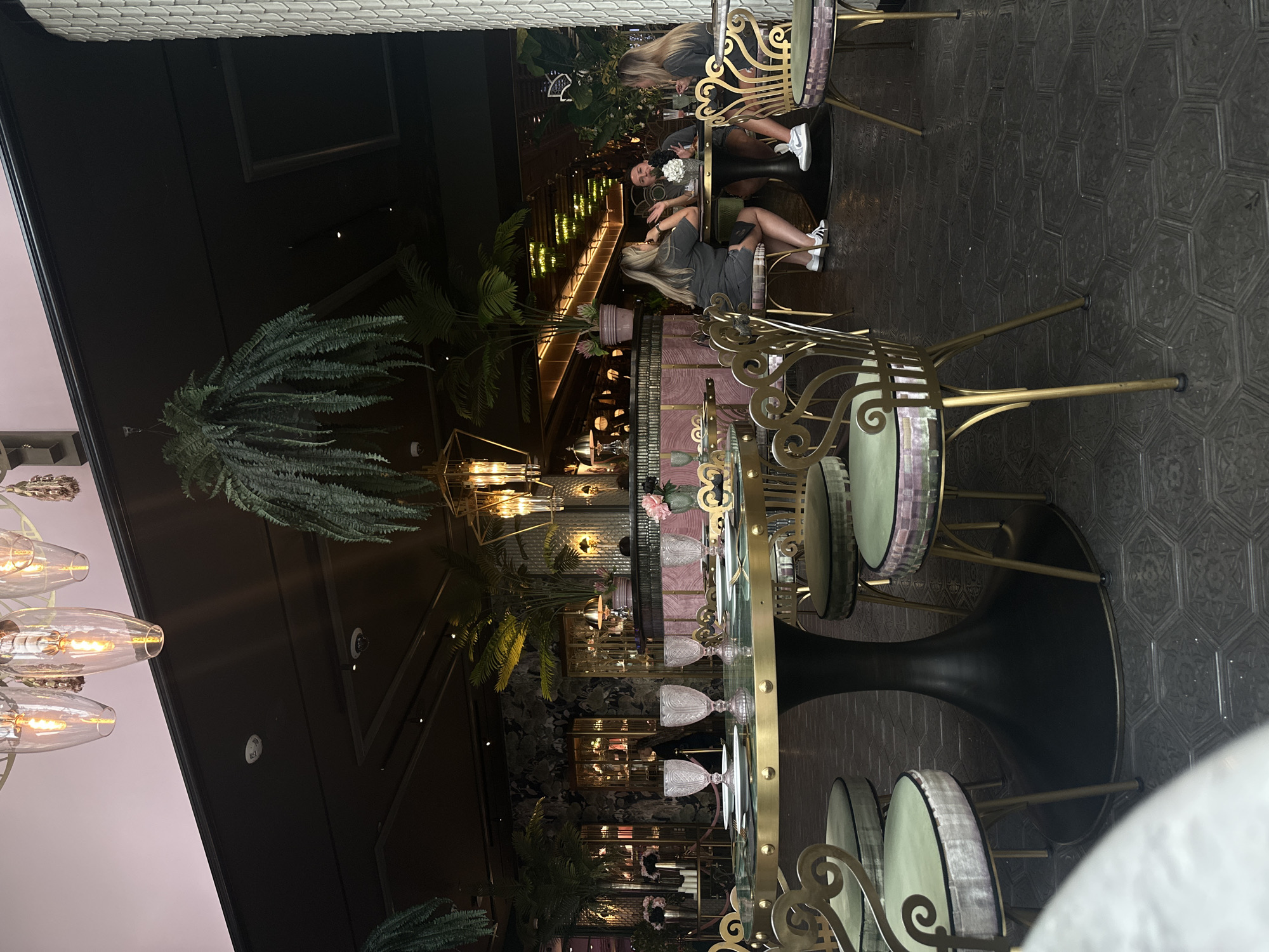 The image depicts an indoor scene of a restaurant in Paradise, United States of America, with a focus on dining tables and patrons. In the foreground, a round table is set with four place settings, each including a white plate and a stemmed glass. The table has a dark, textured base and a gold rim. Several ornate gold chairs with upholstered seats are arranged around this table.
Further back, another round table is occupied by three individuals. Two women and one man are seated, engaged in conversation. They are dressed casually. The table they are seated at also has a dark base and a gold rim. Drinks are visible on this table, along with a small floral arrangement.
The restaurant's interior features lush greenery, including hanging ferns and potted plants, contributing to an upscale botanical ambiance. The decor includes gold accents, such as the chairs and table rims, and is illuminated by decorative pendant lights. The walls appear to be a deep green or black, with some sections featuring textured wallpaper or paneling. A bar area with pink upholstery is visible in the mid-ground. The flooring is a dark, hexagonal tile with an intricate pattern. The overall lighting suggests an evening or dimly lit setting.