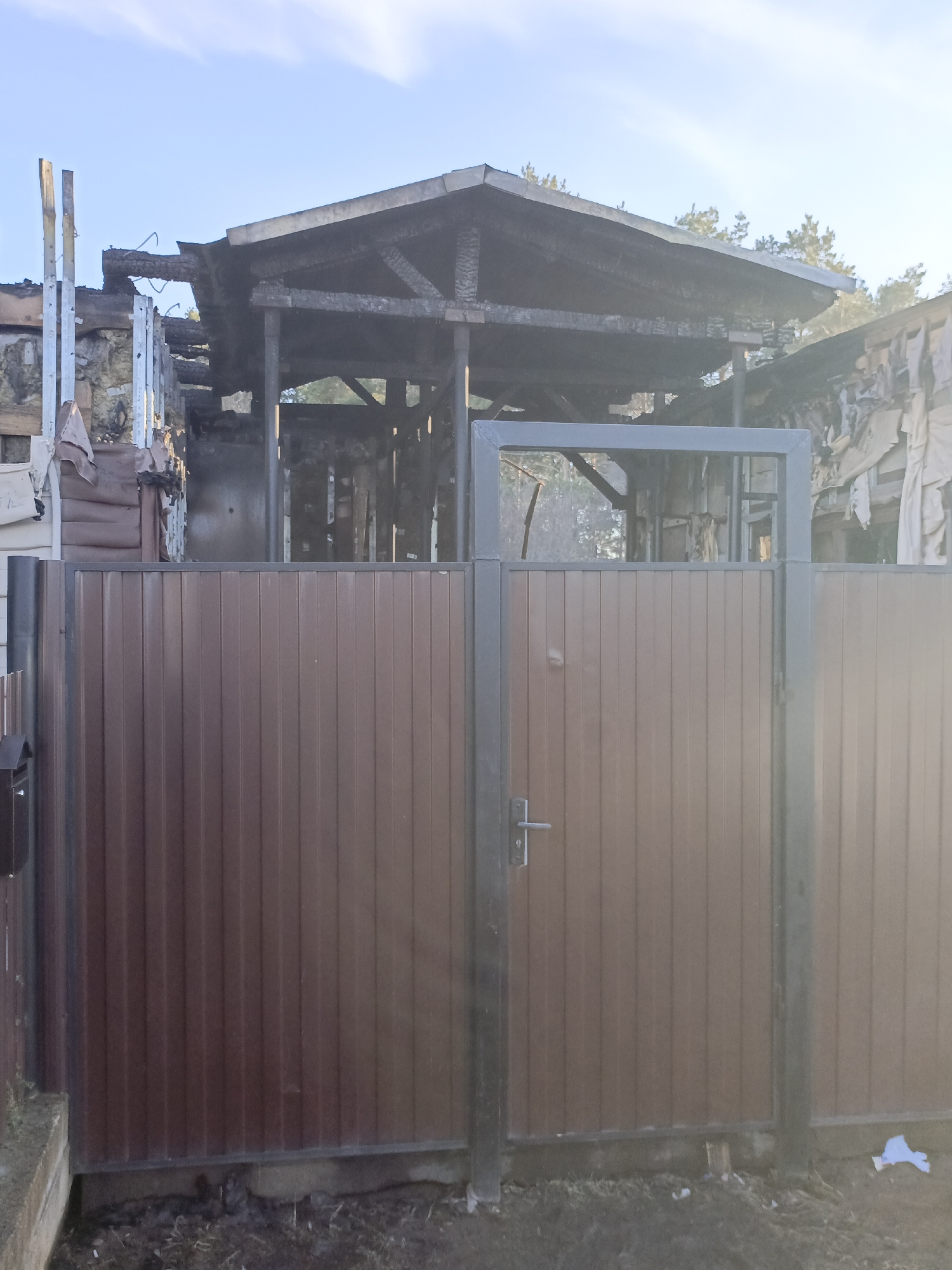 The image shows the aftermath of a fire that has destroyed a building located in Chaikovsky, Russia. A metal fence partially obscures the burned structure. The remaining wooden framework of the building is charred. The roof is mostly gone, with remnants of burned wood and beams visible. The fence is constructed of metal posts and brown metal panels. There is evidence of fire damage on structures adjacent to the main building. The sky is pale blue with sunlight. There is debris on the ground.
