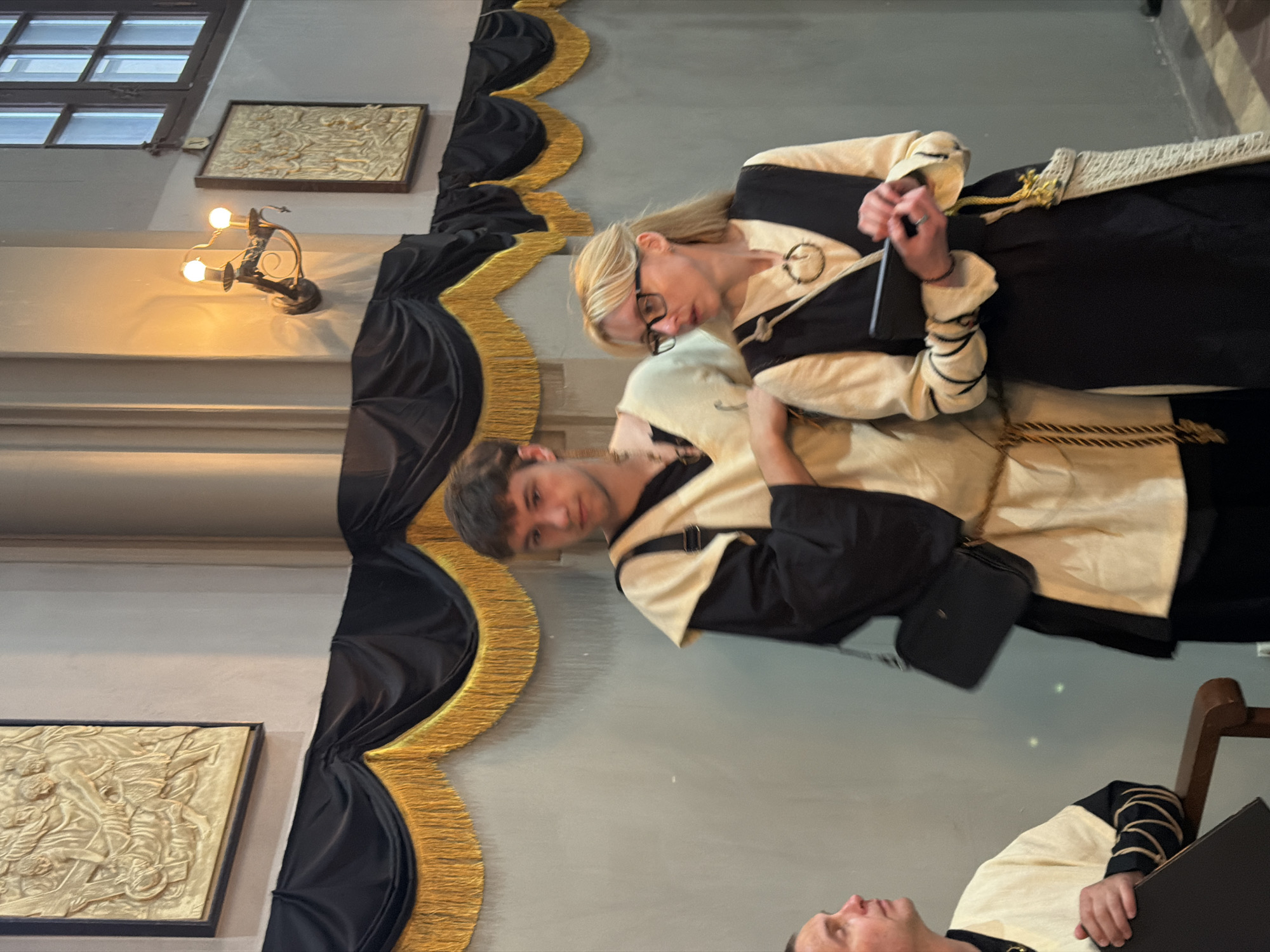 The image, when viewed upright, depicts an indoor scene featuring three individuals dressed in historical or period-style attire. The background consists of a light-colored wall decorated with elaborate black fabric drapes featuring gold scalloped trim, suggesting a formal event or display. Two framed, golden bas-reliefs, depicting intricate historical or mythological scenes, are mounted on the wall. A wall-mounted sconce with two exposed light bulbs provides warm, artificial illumination. A multi-paned window is visible in the upper part of the wall.
In the center, a woman with blonde hair and glasses wears a cream and black tunic-like outfit adorned with a circular brooch. She is holding a dark rectangular object, possibly a phone or small book, and appears focused. Next to her, a young man with dark hair, dressed in a similar cream and black tunic, looks towards the camera with a slight smile. He carries a small black bag over his shoulder. Partially visible in the lower right, a third person, also in a matching costume, holds a dark binder or large book.
The subjects' attire and the decorative setting suggest they are participating in a historical reenactment, a themed event, or working as guides/performers in a significant location, likely a historical building or museum in Saint Petersburg, Russia. The artificial lighting indicates the event could be taking place in the evening or in a purposefully dim environment. There is no visible text in the media.