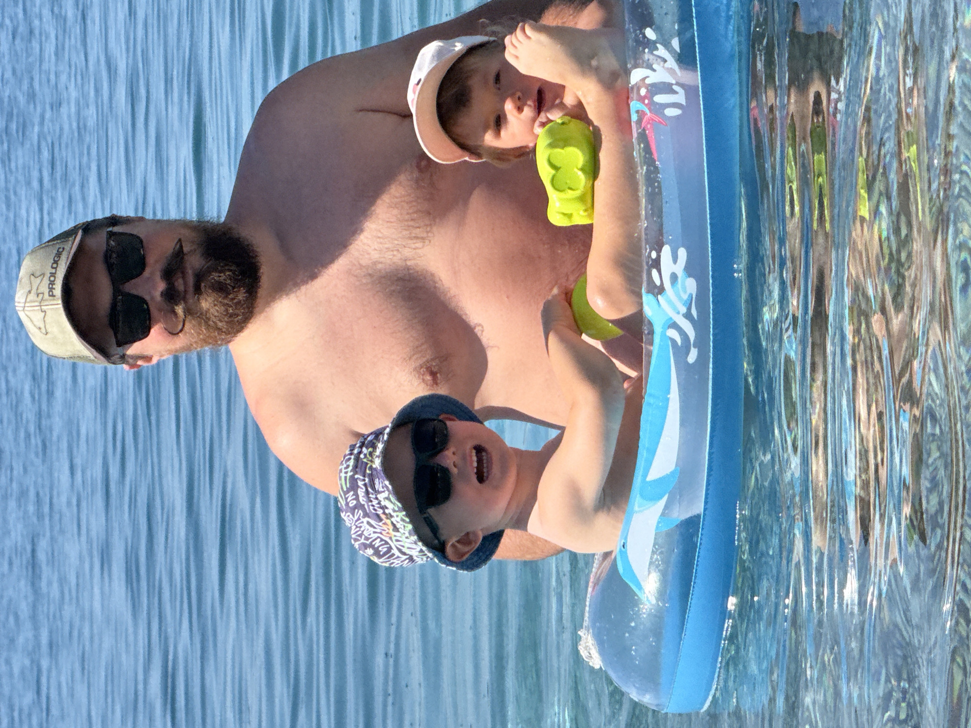 An outdoor scene in Boreti, Montenegro, depicts an adult male and two young children in a clear, blue body of water under sunlight. The adult male, positioned behind the children and partially submerged, is shirtless with a full beard and a styled handlebar mustache. He wears dark sunglasses and a camouflage-patterned baseball cap with "PROLOGIC" visible on the front. In front of him, two children are floating within a blue and clear inflatable ring. The child on the left, a young boy, wears a patterned bucket hat and dark sunglasses, smiling with his mouth open while holding a small yellow object. The child on the right, a young girl, wears a light-colored baseball cap and holds a bright green toy. Both children are looking towards the viewer. The water surface exhibits ripples and reflections of sunlight, with only the open water visible in the background.